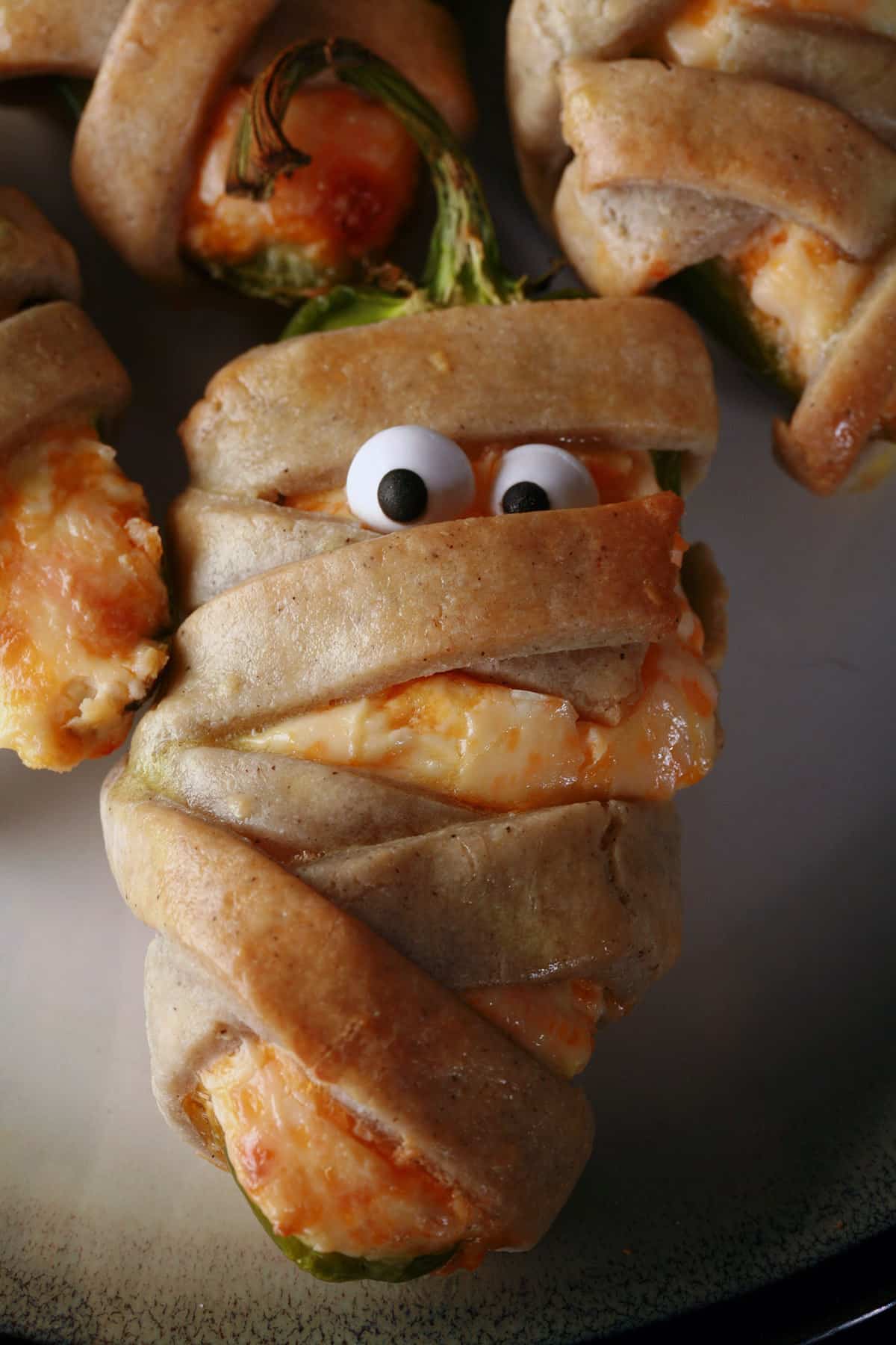 Several gluten free mummy jalapeno poppers on a plate. Each has 2 candy eyeballs.