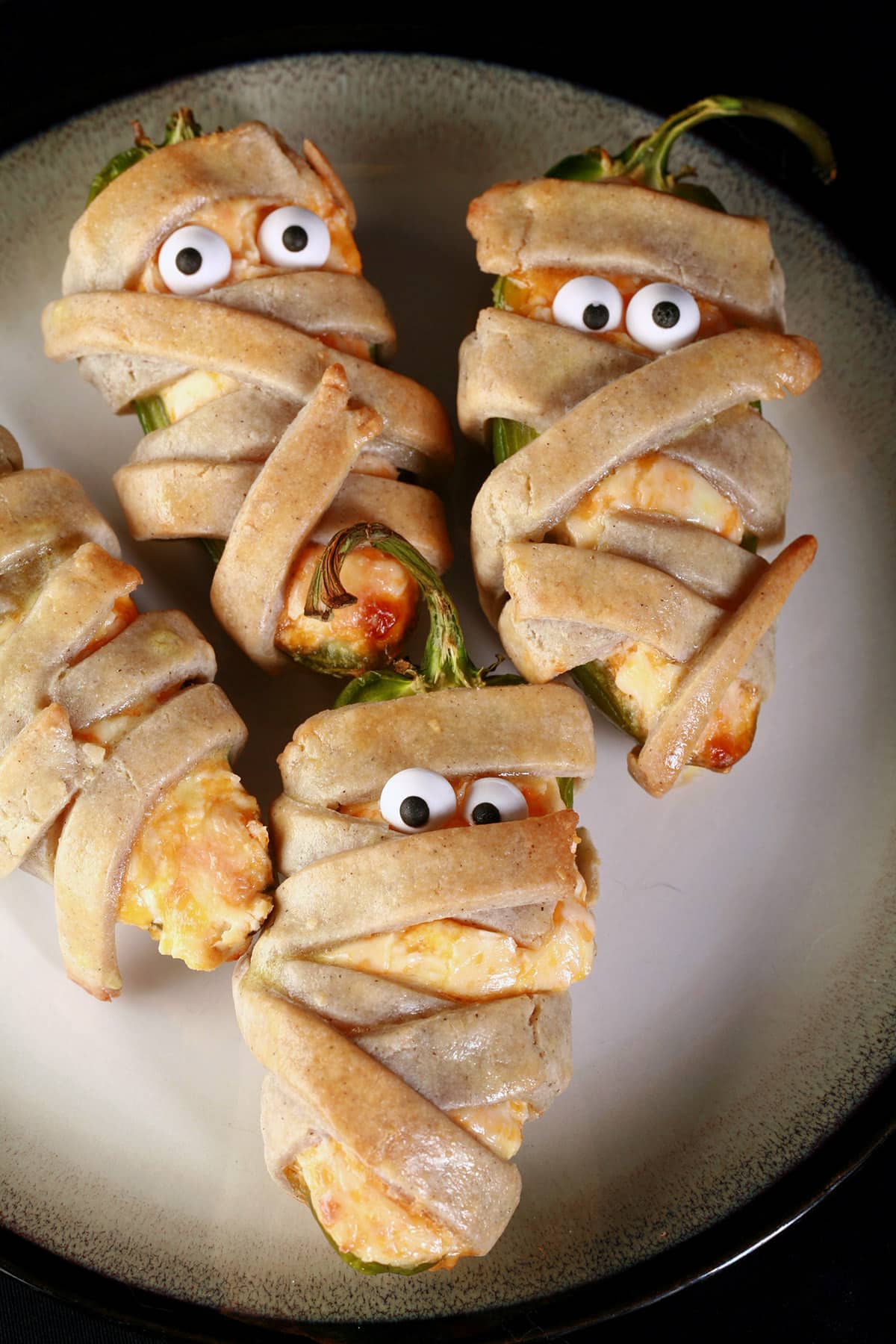 Several gluten free mummy jalapeno poppers on a plate. Each has 2 candy eyeballs.