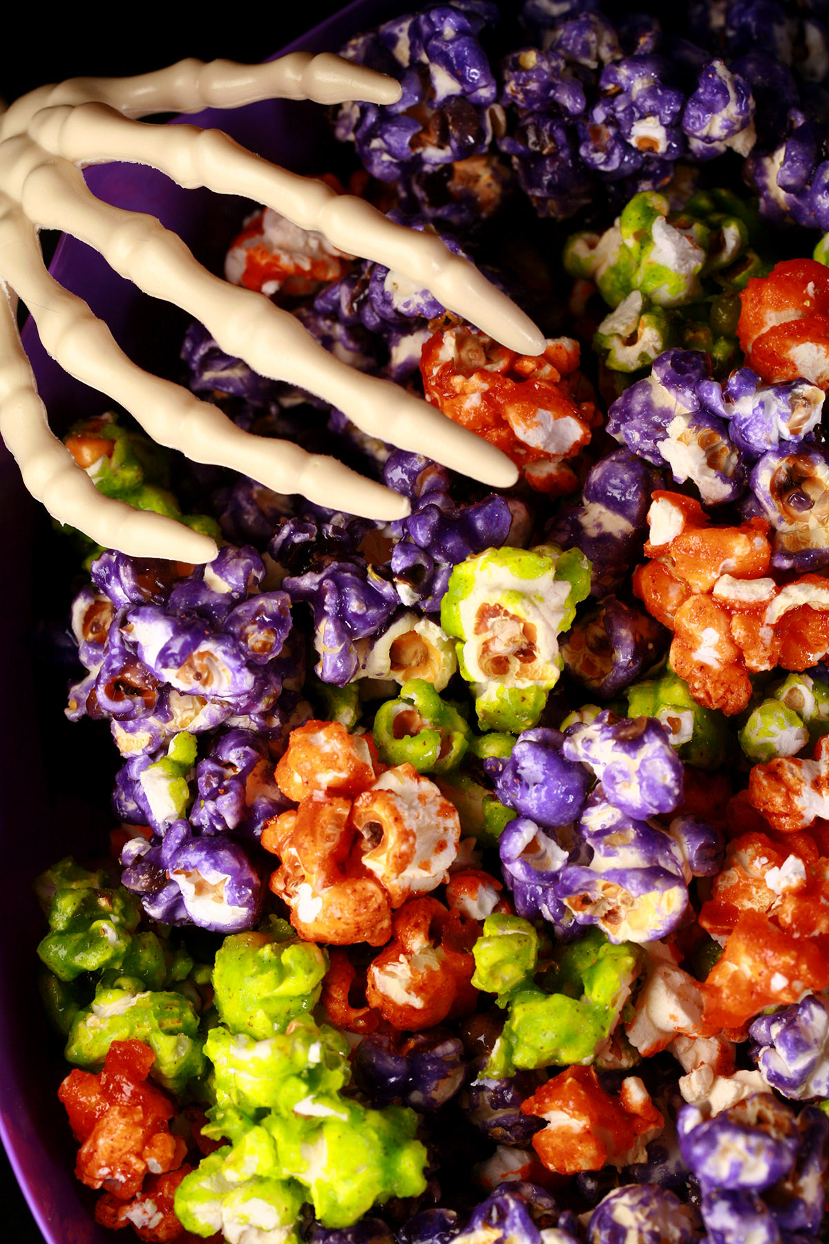 A bowl of Halloween popcorn - orange, purple, and lime green glazed popcorn, mixed together. A pair of skeleton hand serving spoons are visible.