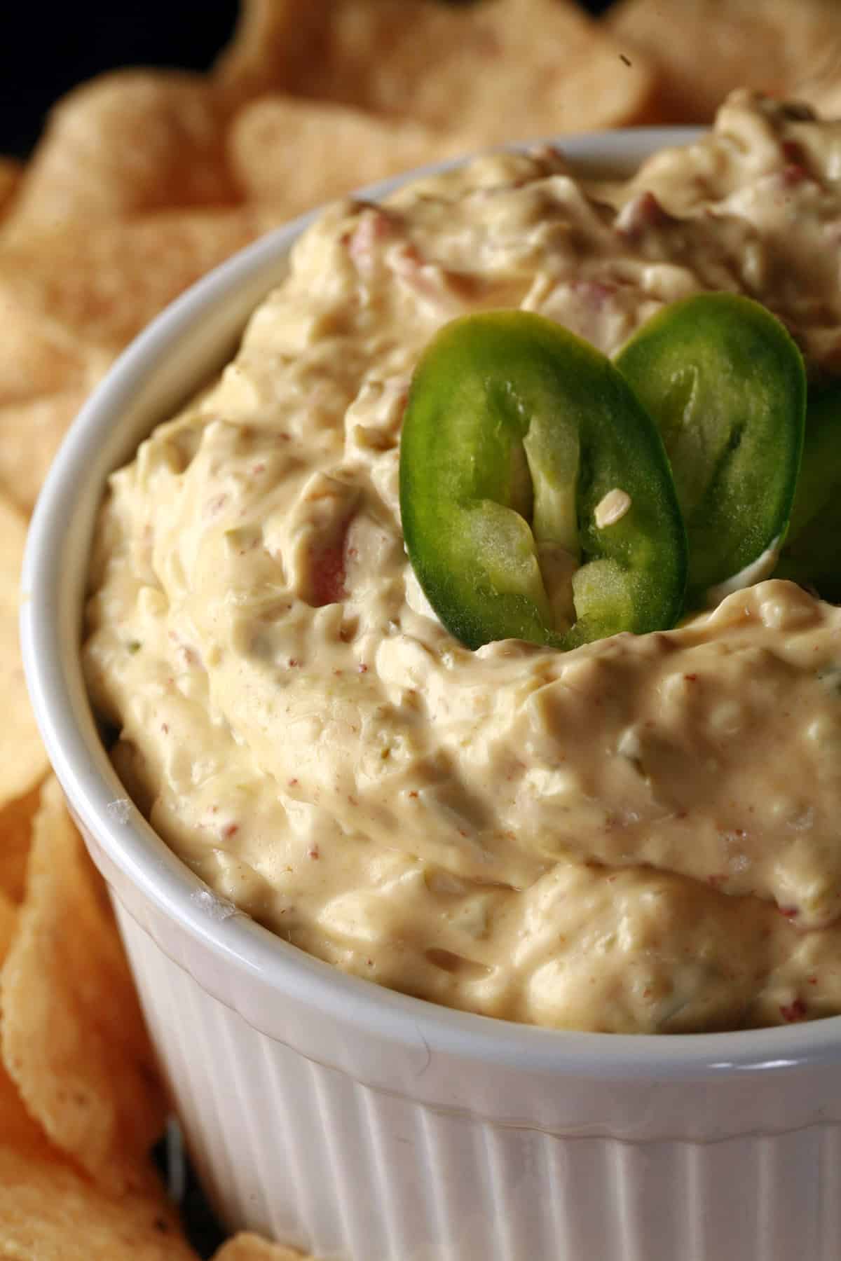 A ramekin of bacon jalapeno dip, surrounded by corn chips and topped with sliced jalapenos.