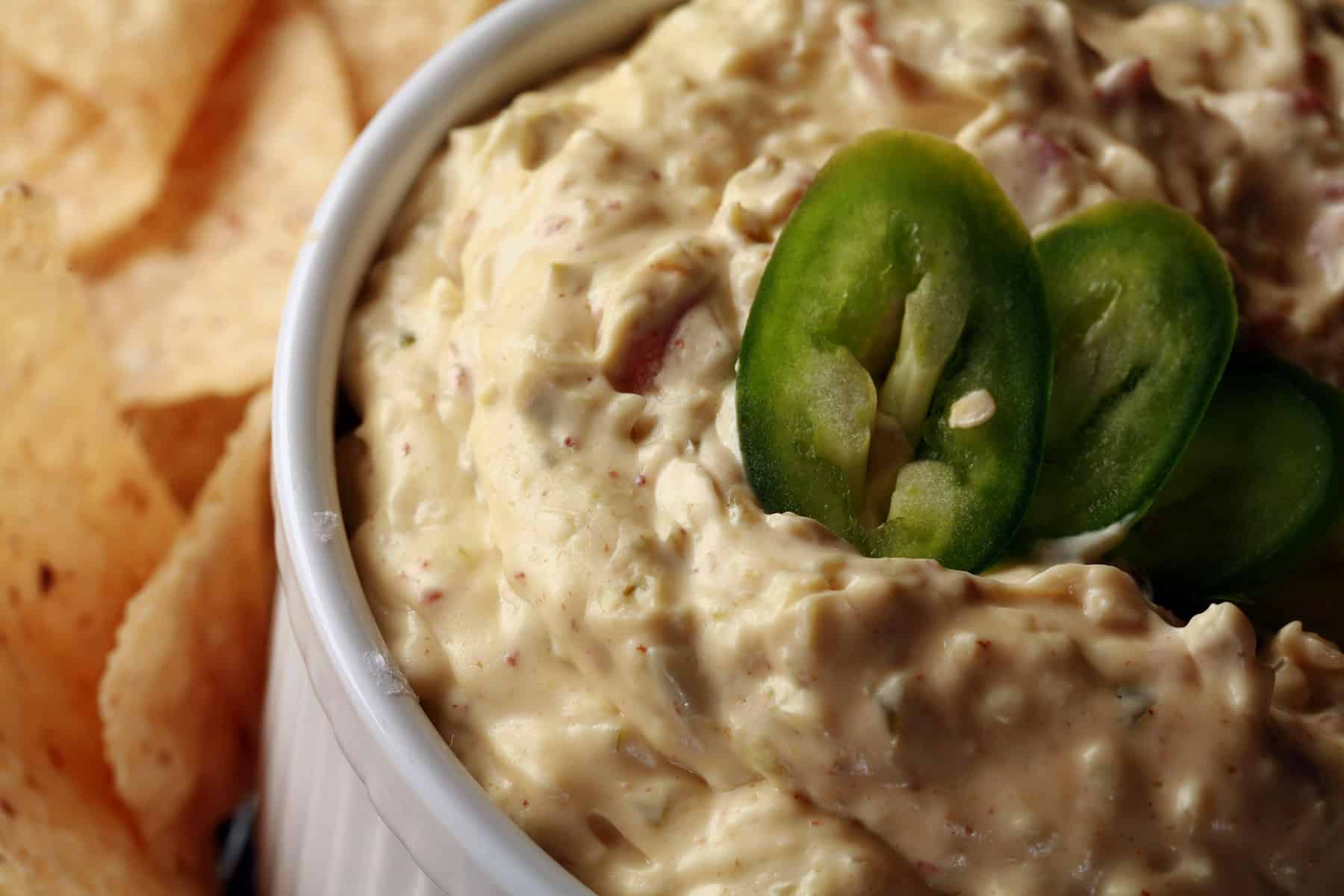 A ramekin of gluten free jalapeno dip, surrounded by corn chips and topped with sliced jalapenos.