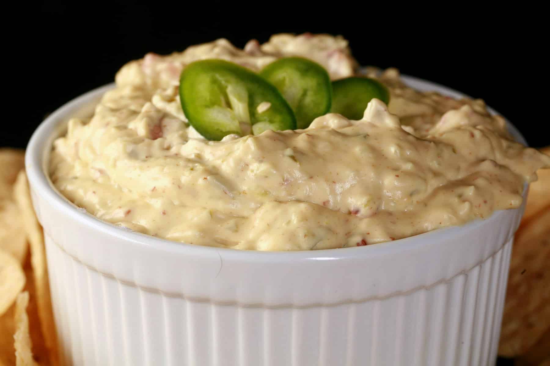 A ramekin of bacon jalapeno dip, surrounded by corn chips and topped with sliced jalapenos.