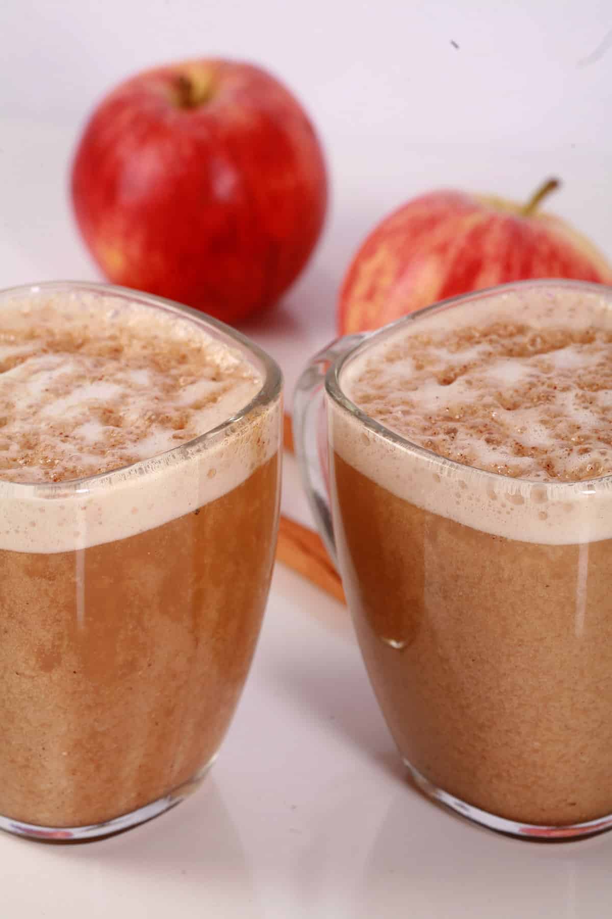 2 glass mugs of apple crisp oatmilk macchiato, with cinnamon sticks and apples next to them.