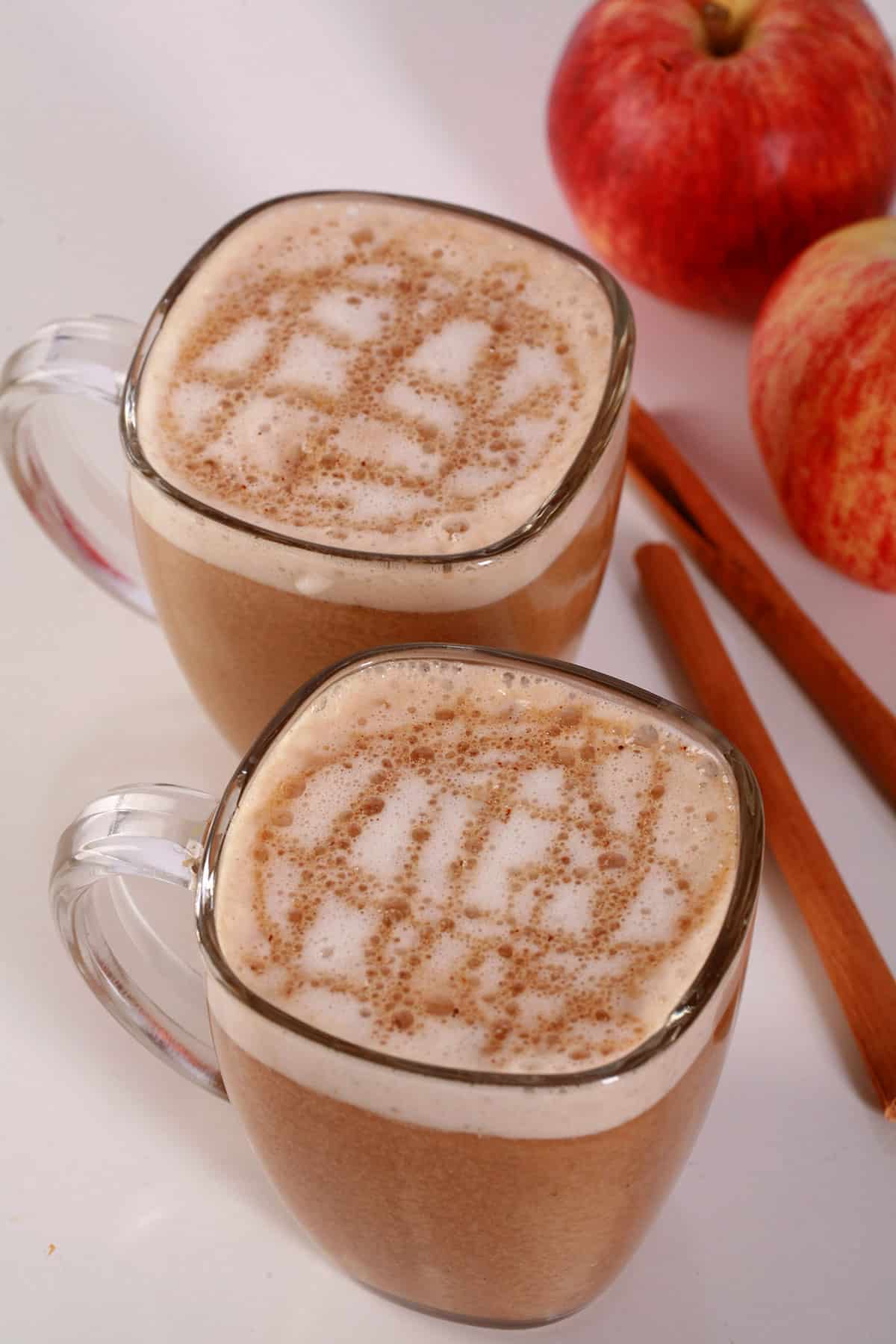 2 glass mugs of apple crisp oatmilk macchiato, with cinnamon sticks and apples next to them.