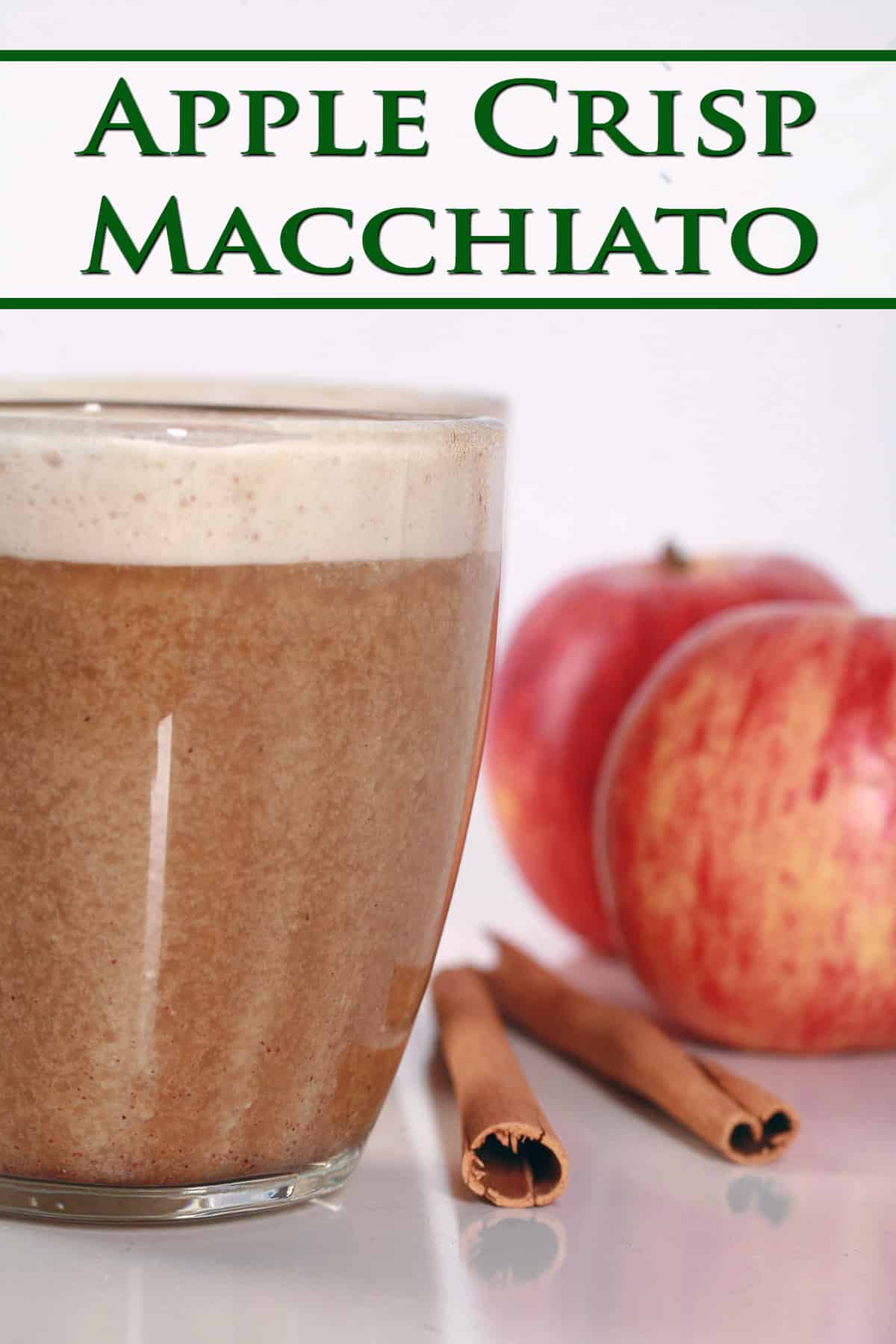 A glass mug of apple crisp macchiato, with cinnamon sticks and apples next to it.