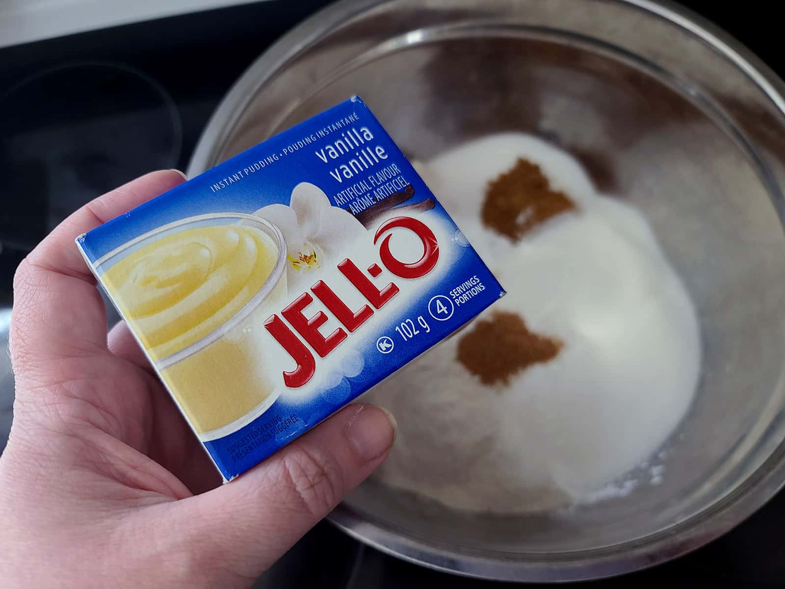 A hand holds a box of Jello vanilla pudding mix over the bowl of dry ingredients.