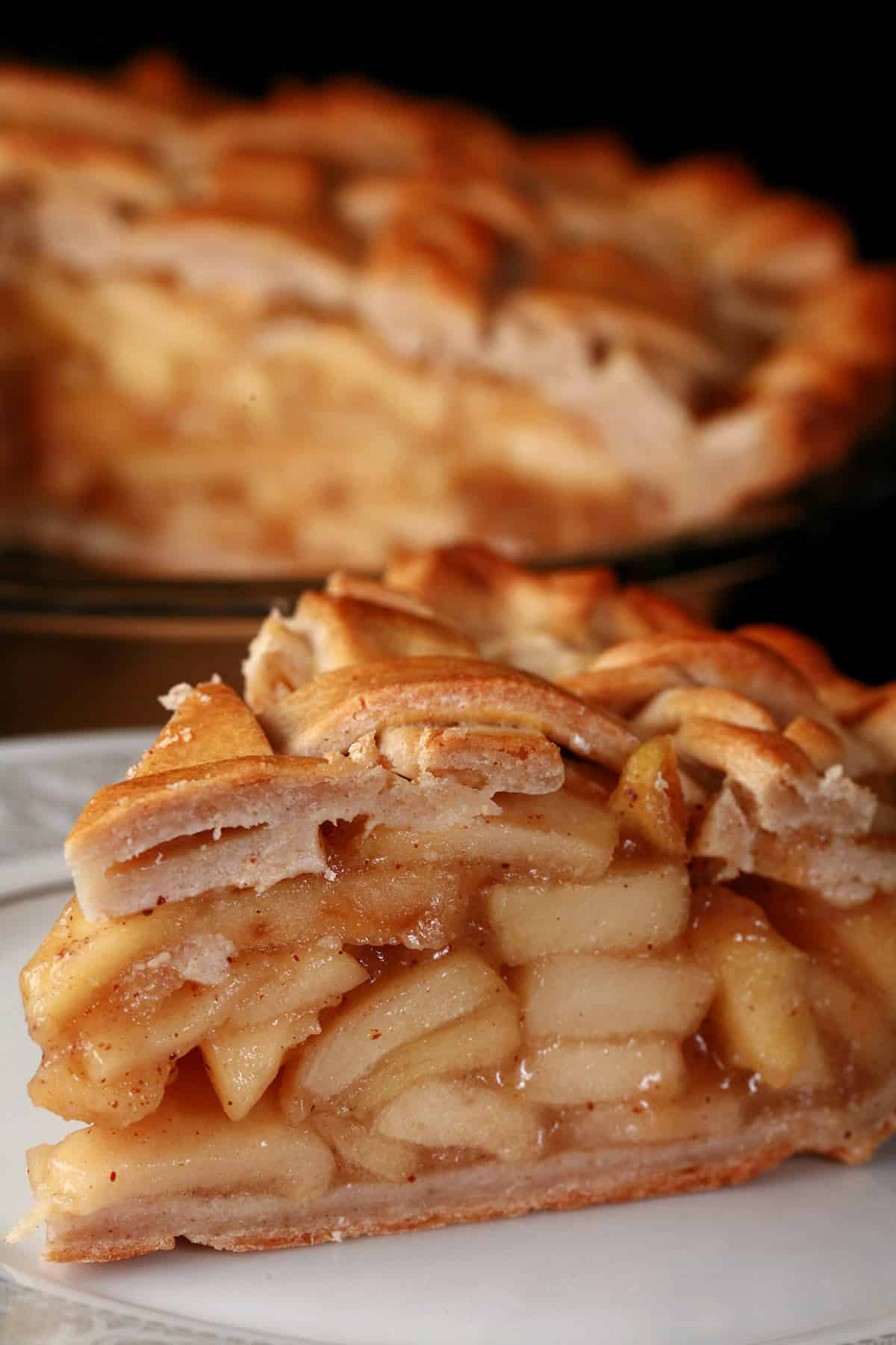 A slice of lattice top gluten-free apple pie on a plate, in front of the rest of the pie.