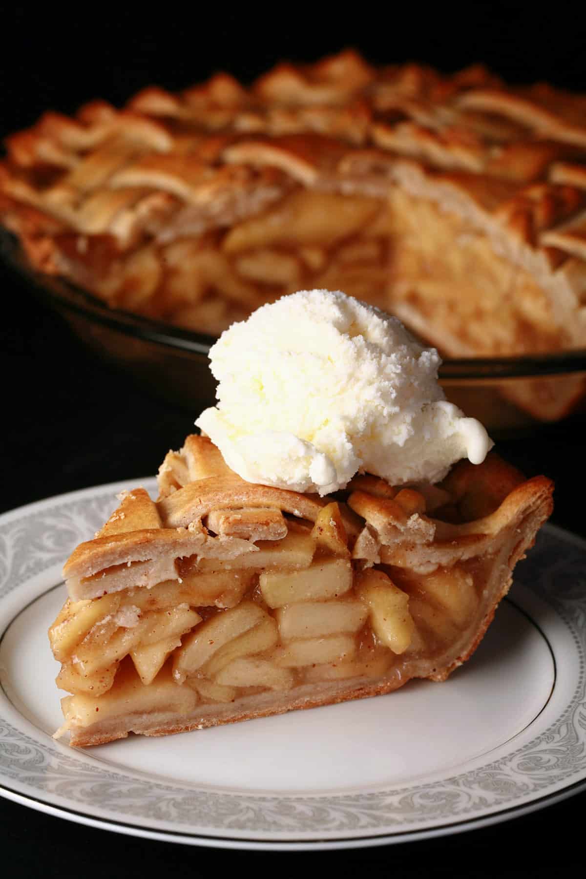 A slice of lattice top gluten free apple pie on a plate, in front of the rest of the pie.