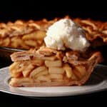 A slice of lattice top gluten-free apple pie on a plate, in front of the rest of the pie.