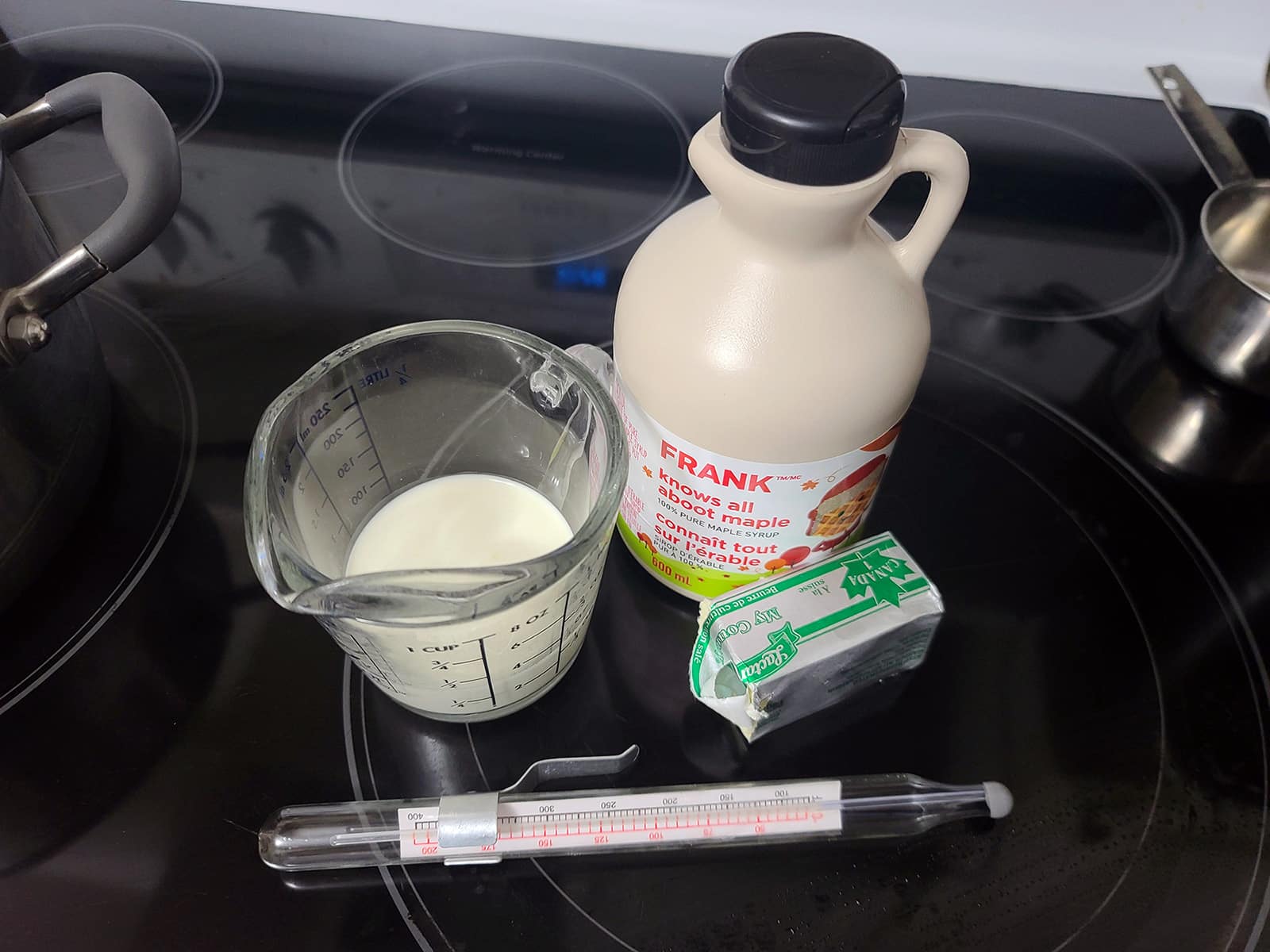 A jug of maple syrup, a chunk of butter, a glass measuring cup with heavy cream, and a candy thermometer.