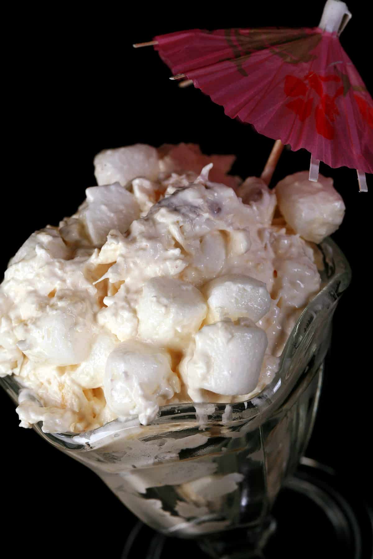 Pina colada marshmallow salad in a tulip glass with a pink cocktail umbrella sticking out.