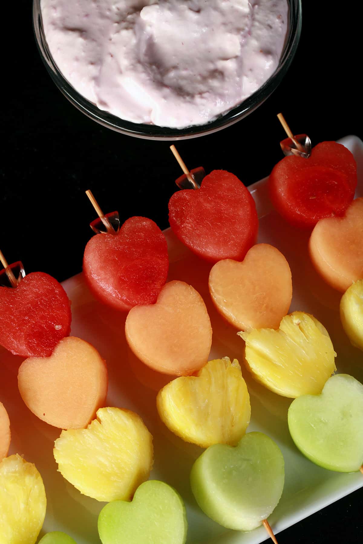 Several heart shaped fruit skewers- watermelon, cantaloupe, pineapple, and honeydew - next to a bowl of raspberry cheesecake dip.