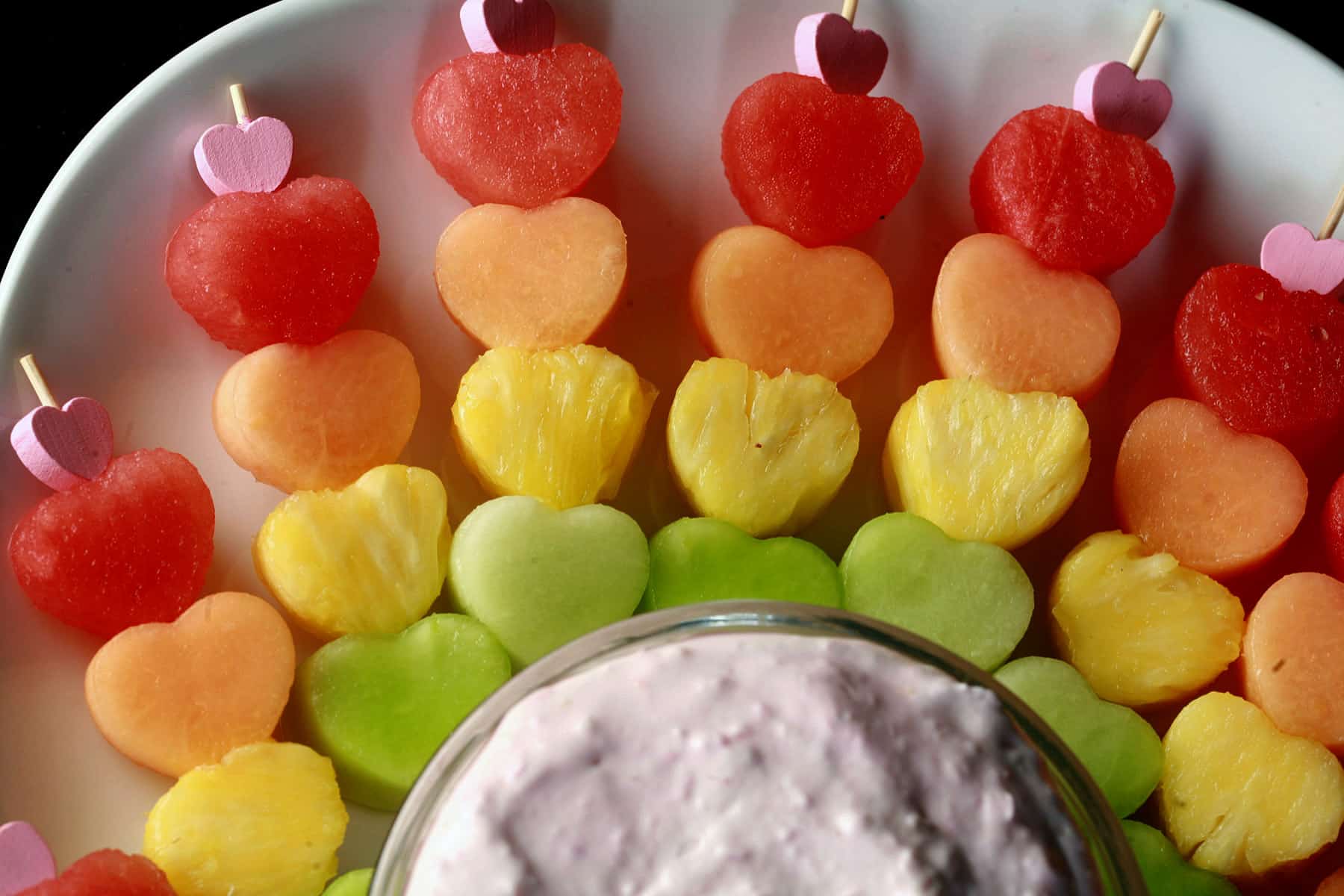 Several heart shaped fruit skewers- watermelon, cantaloupe, pineapple, and honeydew - next to a bowl of raspberry cheesecake dip.