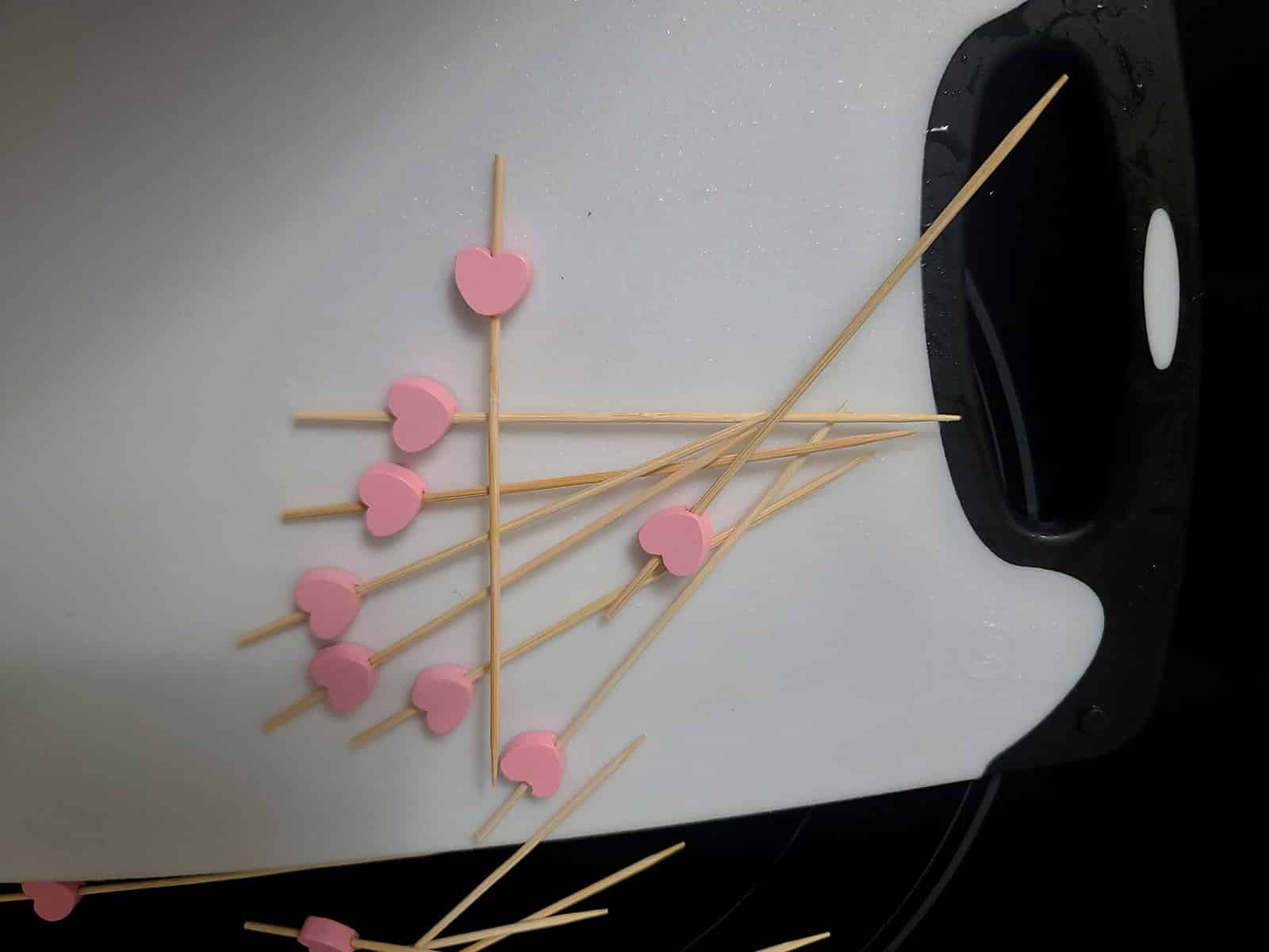 Several skewers with a pink heart decoration on the end, spread on a white cutting board.
