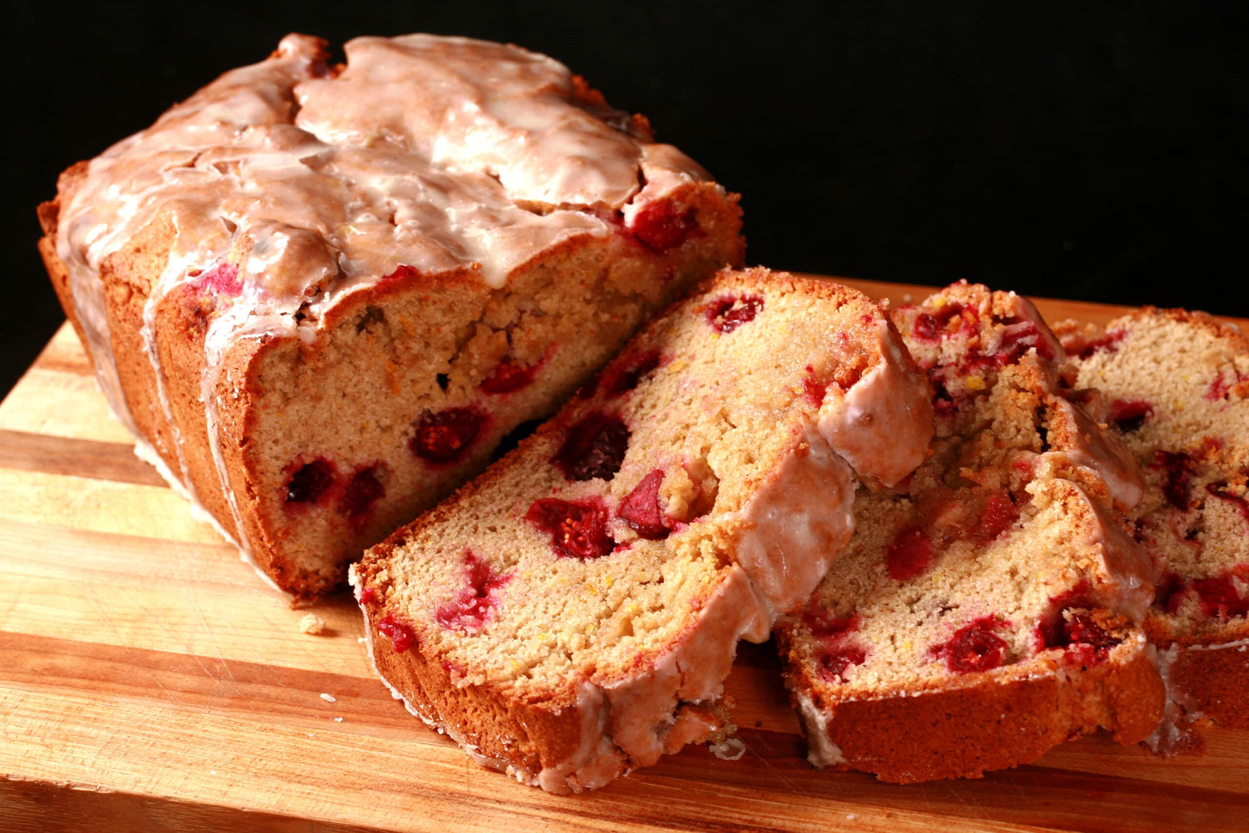 A loaf of glazed gluten-free cranberry orange bread, sliced on a cutting board.