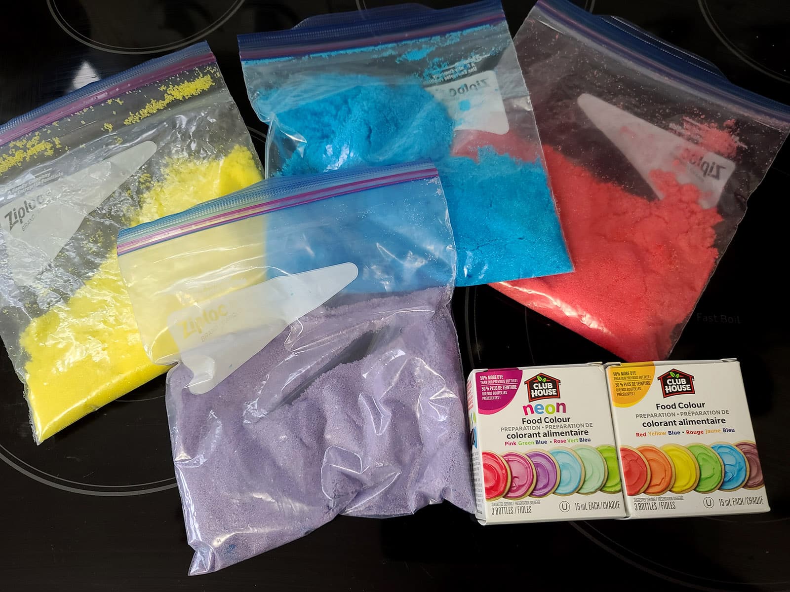 Bags of pink, purple, blue, and yellow colored sugar, along with a box of liquid food coloring.