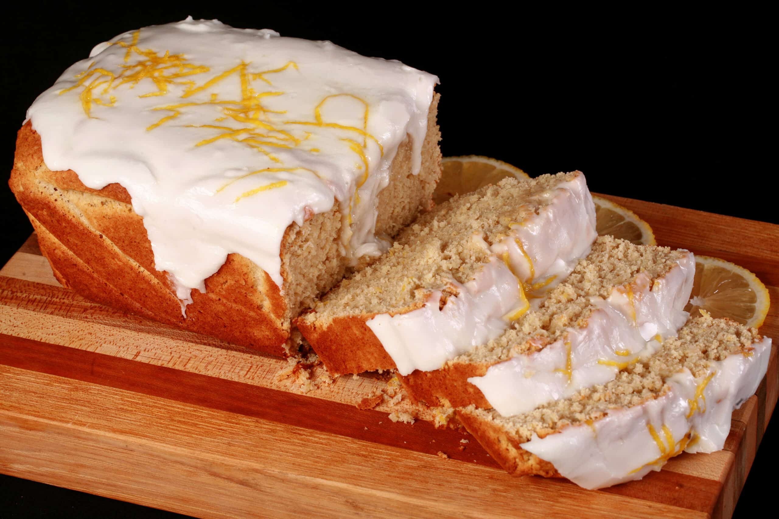 A sliced loaf of gluten free lemon drizzle cake, with white glaze and lemon zest on top.
