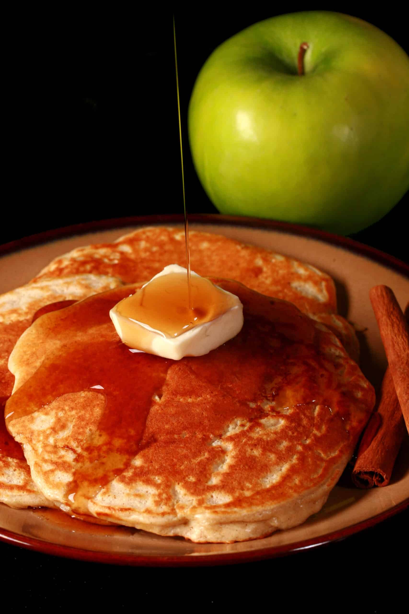 3 apple cinnamon protein pancakes on a plate, topped with butter and maple syrup.  There are 2 cinnamon sticks on the plate, and a green apple beside it..