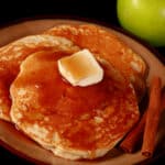 3 apple cinnamon protein pancakes on a plate, topped with butter and maple syrup. There are 2 cinnamon sticks on the plate, and a green apple beside it..