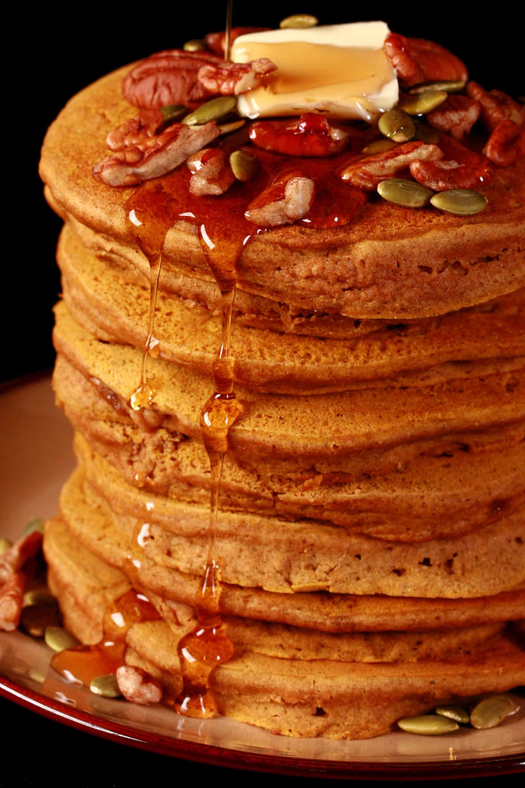 A tall stack of gluten free pumpkin pancakes, topped with butter, maple syrup, pecans, and pumpkin seeds.