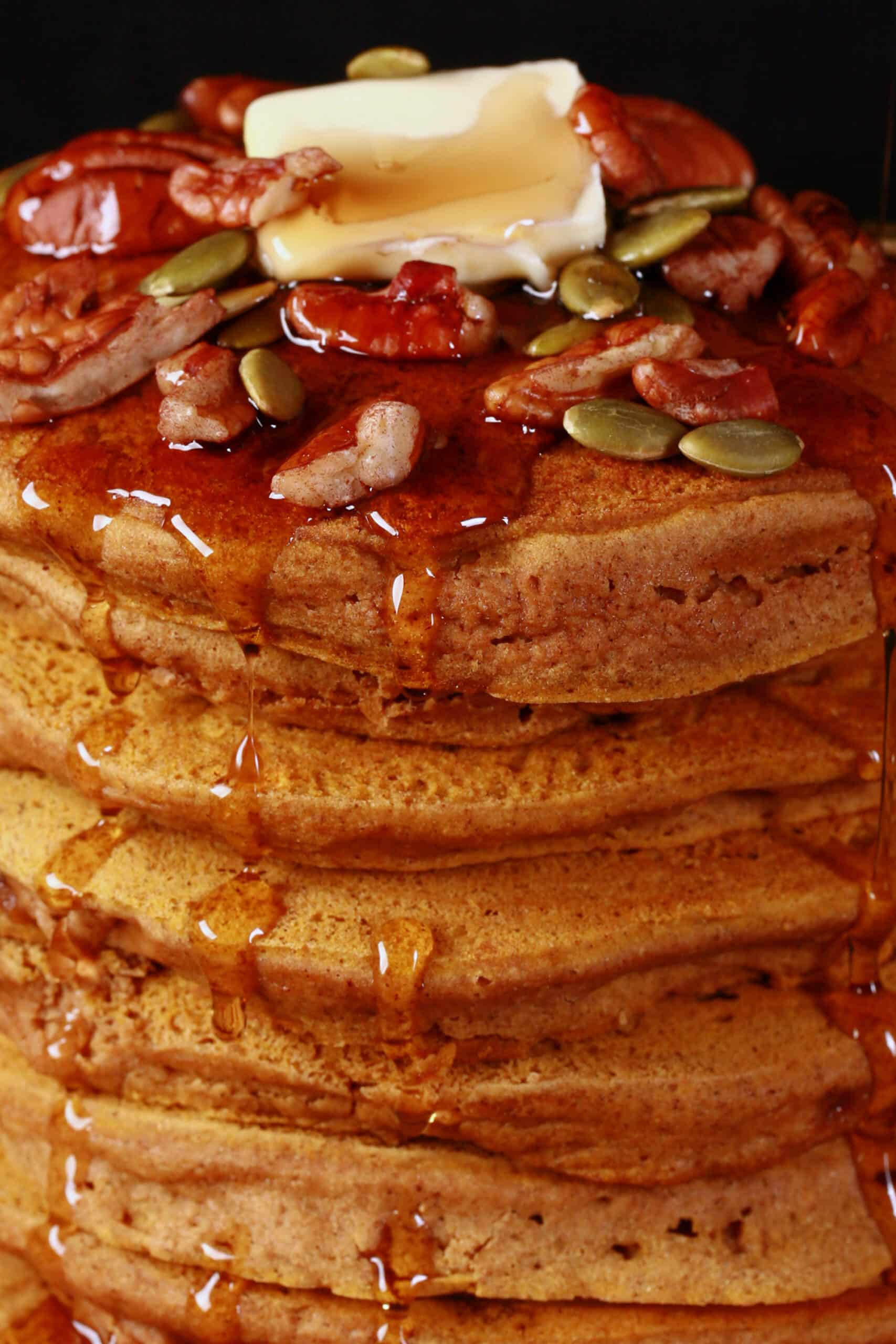 A tall stack of gluten-free pumpkin pancakes, topped with butter, maple syrup, pecans, and pumpkin seeds.