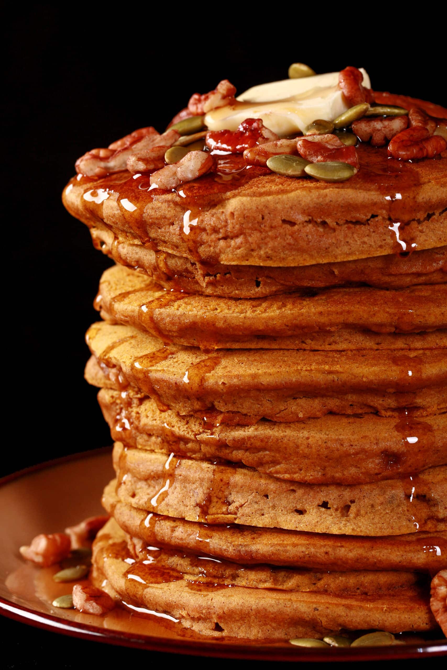 A tall stack of gluten free pumpkin pancakes, topped with butter, maple syrup, pecans, and pumpkin seeds.