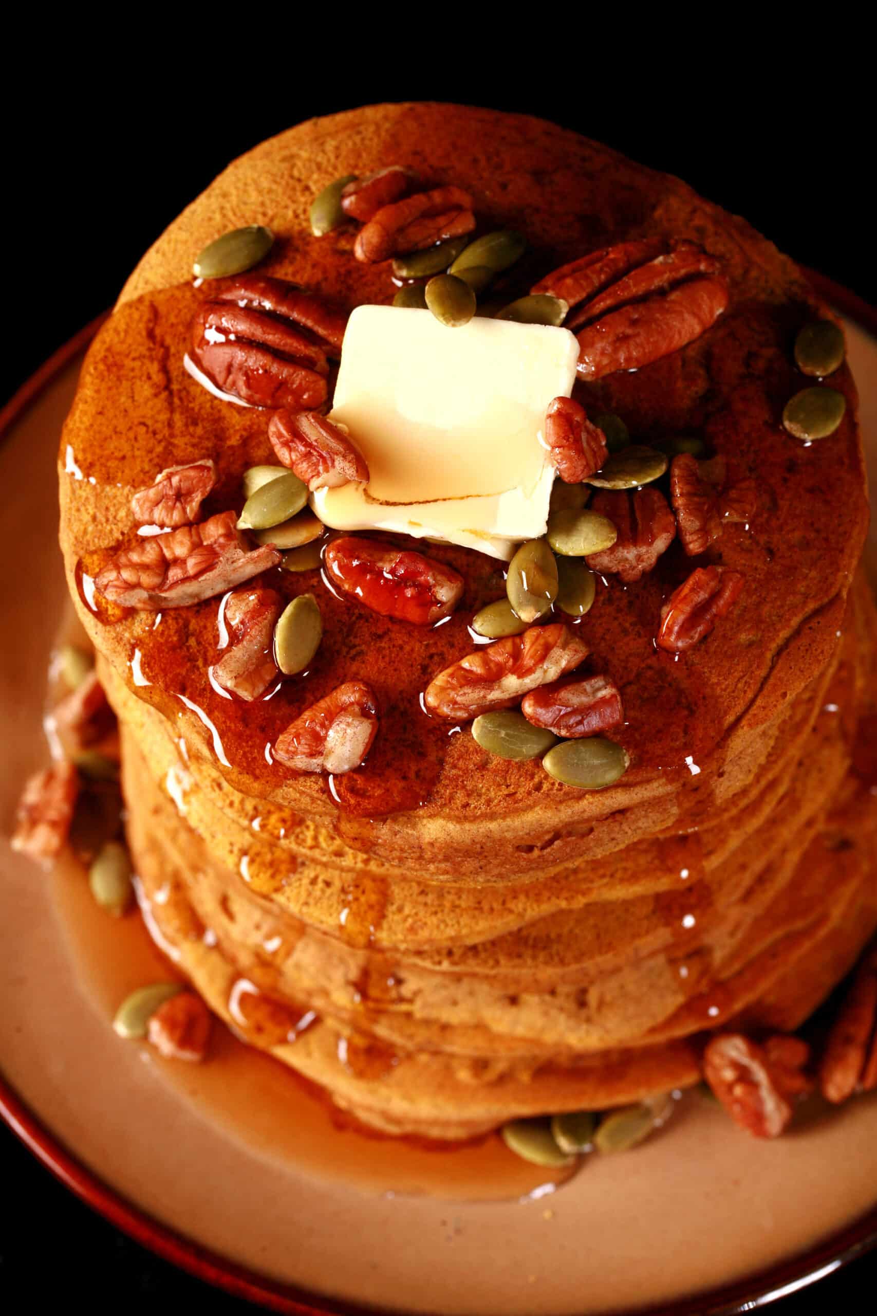 A tall stack of gluten-free pumpkin pancakes, topped with butter, maple syrup, pecans, and pumpkin seeds.