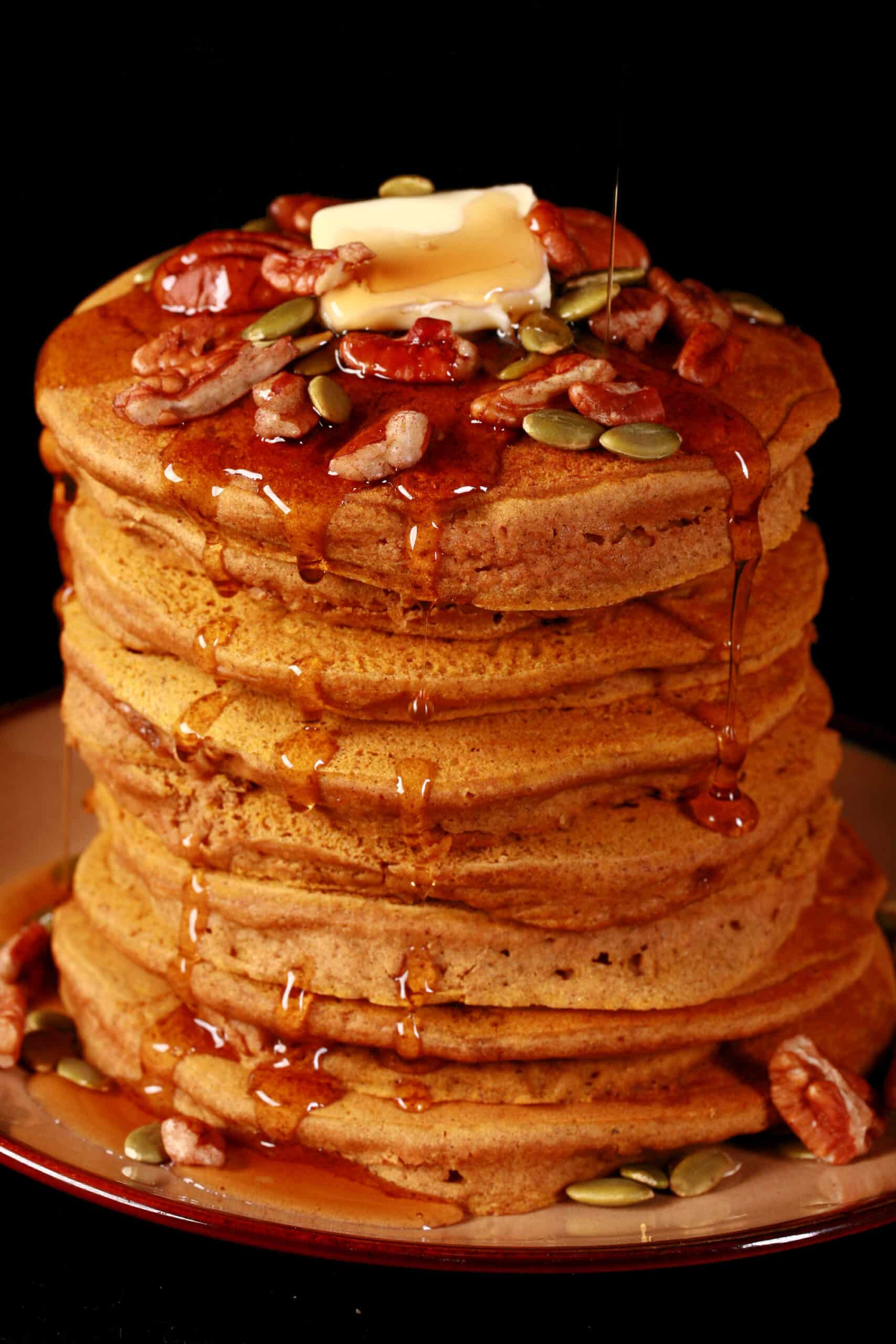 A tall stack of gluten free pumpkin pancakes, topped with butter, maple syrup, pecans, and pumpkin seeds.