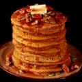 A tall stack of gluten-free pumpkin pancakes, topped with butter, maple syrup, pecans, and pumpkin seeds.