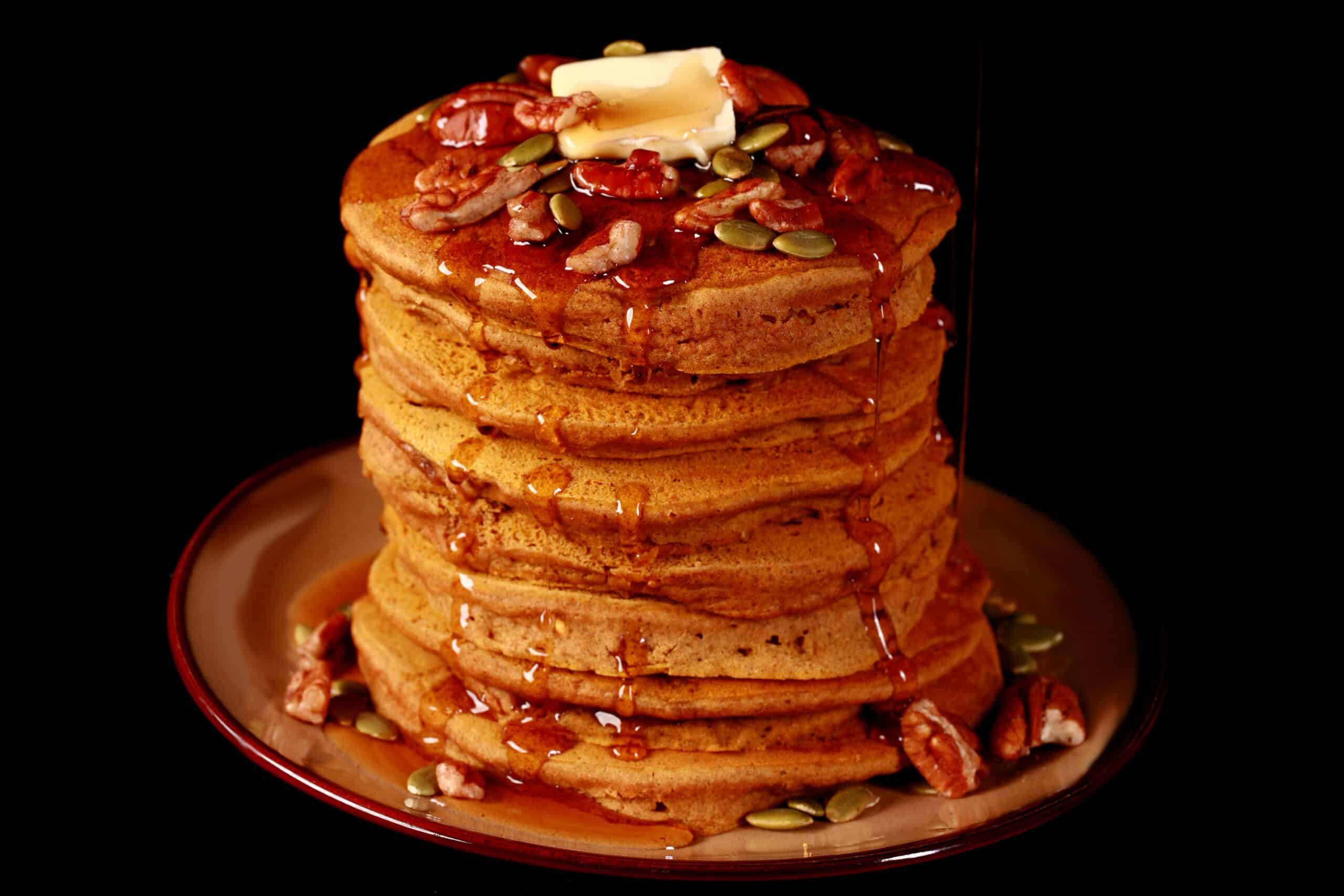 A tall stack of gluten-free pumpkin pancakes, topped with butter, maple syrup, pecans, and pumpkin seeds.