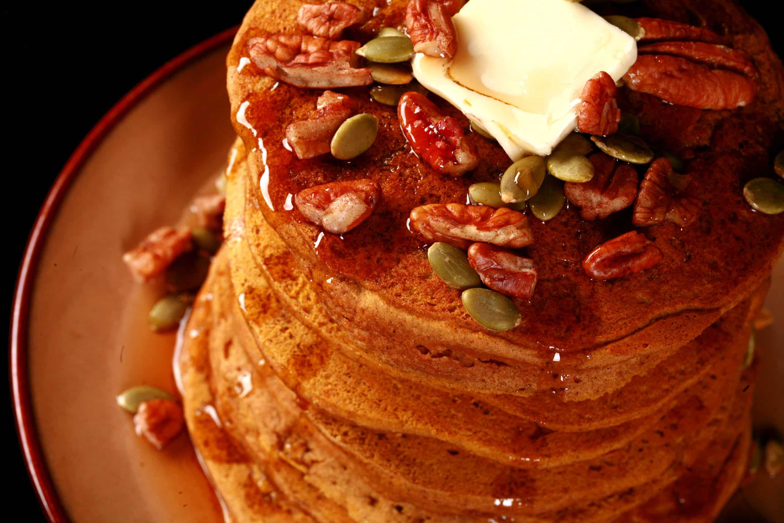 A tall stack of gluten free pumpkin pancakes, topped with butter, maple syrup, pecans, and pumpkin seeds.