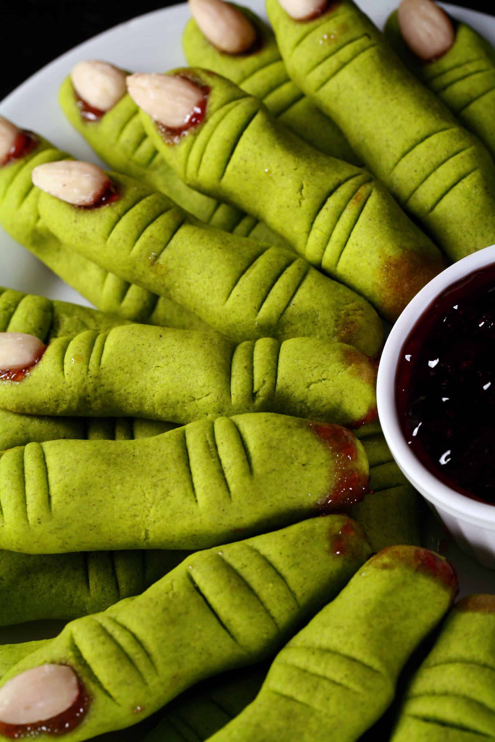 A plate of bright green gluten free witch finger cookies. A plate of bright green gluten free witch finger cookies.  There’s a bowl of red jam next to it, as dip.
