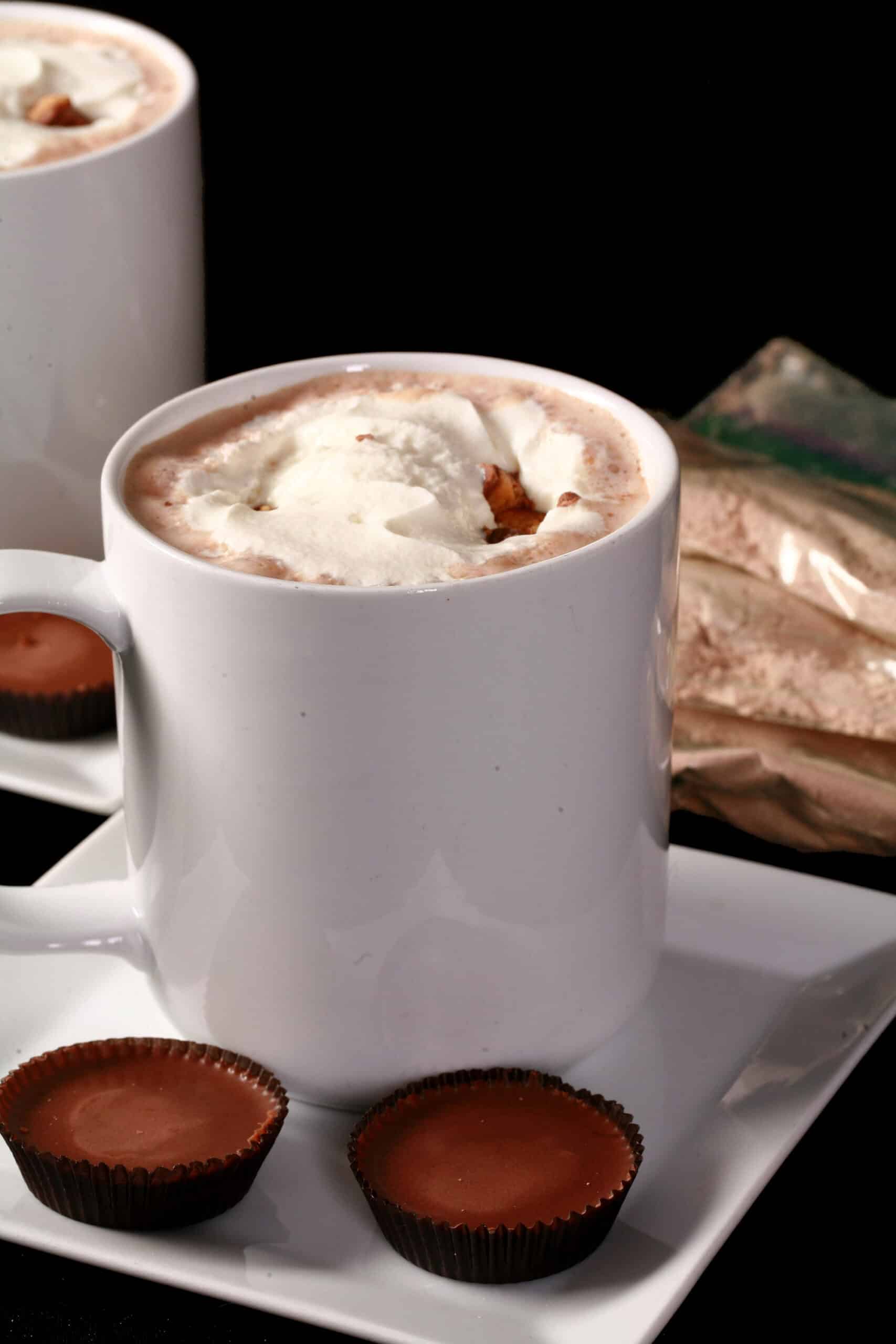 2 mugs of protein peanut butter hot chocolate with whipped cream and chopped peanut butter cups. Several packets of hot chocolate mix are in the background.