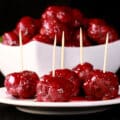 A bowl of gluten free turkey meatballs in a bright red cranberry glaze. There are several meatballs with toothpicks on them on a plate in front of the bowl.