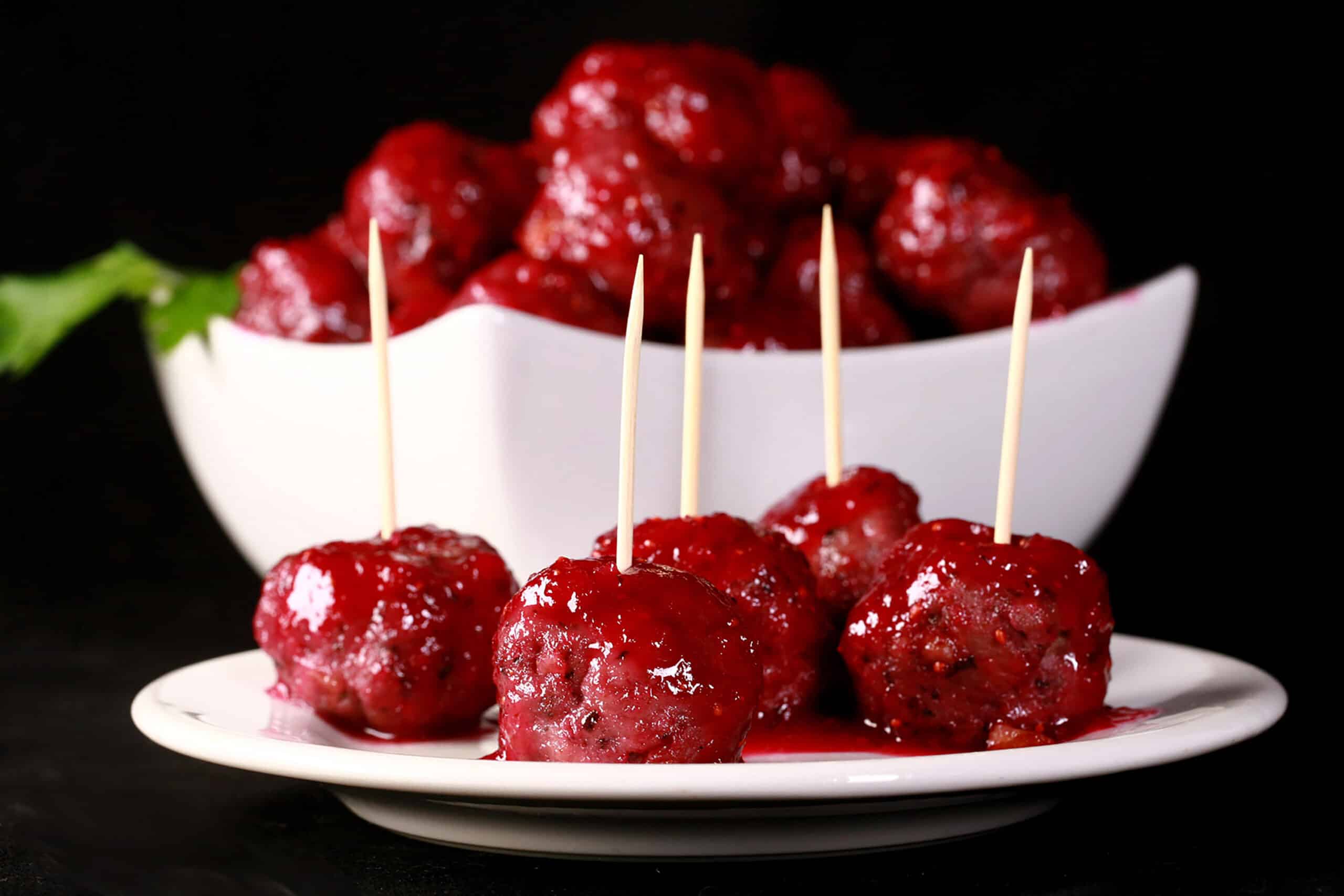 A bowl of gluten free turkey meatballs in a bright red cranberry glaze.  There are several meatballs with toothpicks on them on a plate in front of the bowl.