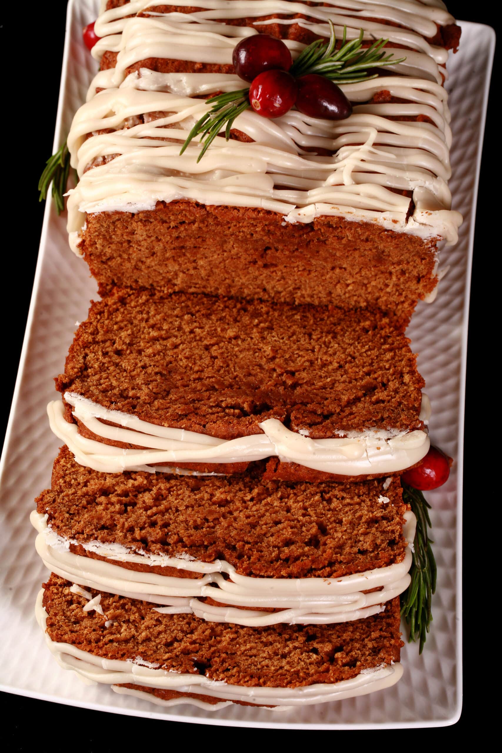 A sliced loaf of gluten-free gingerbread loaf, drizzled with vanilla glaze and garnished with rosemary and cranberries.