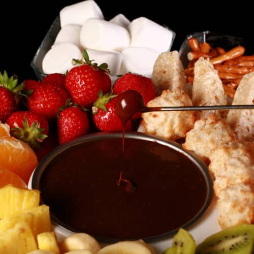 A strawberry being dipped into chocolate fondue, surrounded by pineapple chunks, banana slices, kiwi slices, coconut macaroons, oranges, pretzels, marshmallows, and more.