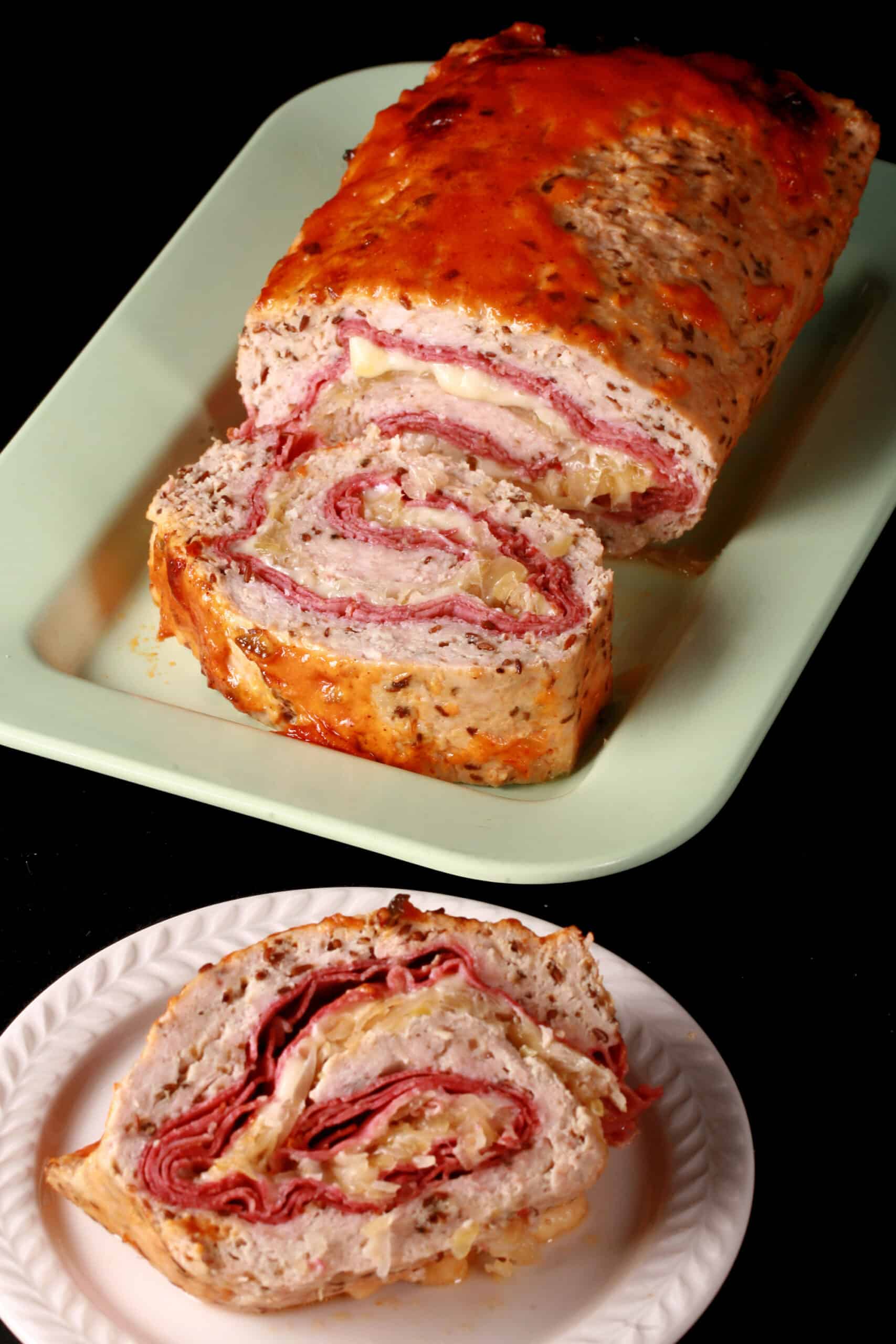 A slice of Reuben Stuffed Meatloaf on a plate in front of the rest of the reuben meatloaf.