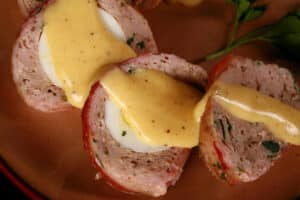 A sliced individual breakfast meatloaf, wrapped in bacon, stuffed with a hard boiled egg, and topped with hollandaise sauce.