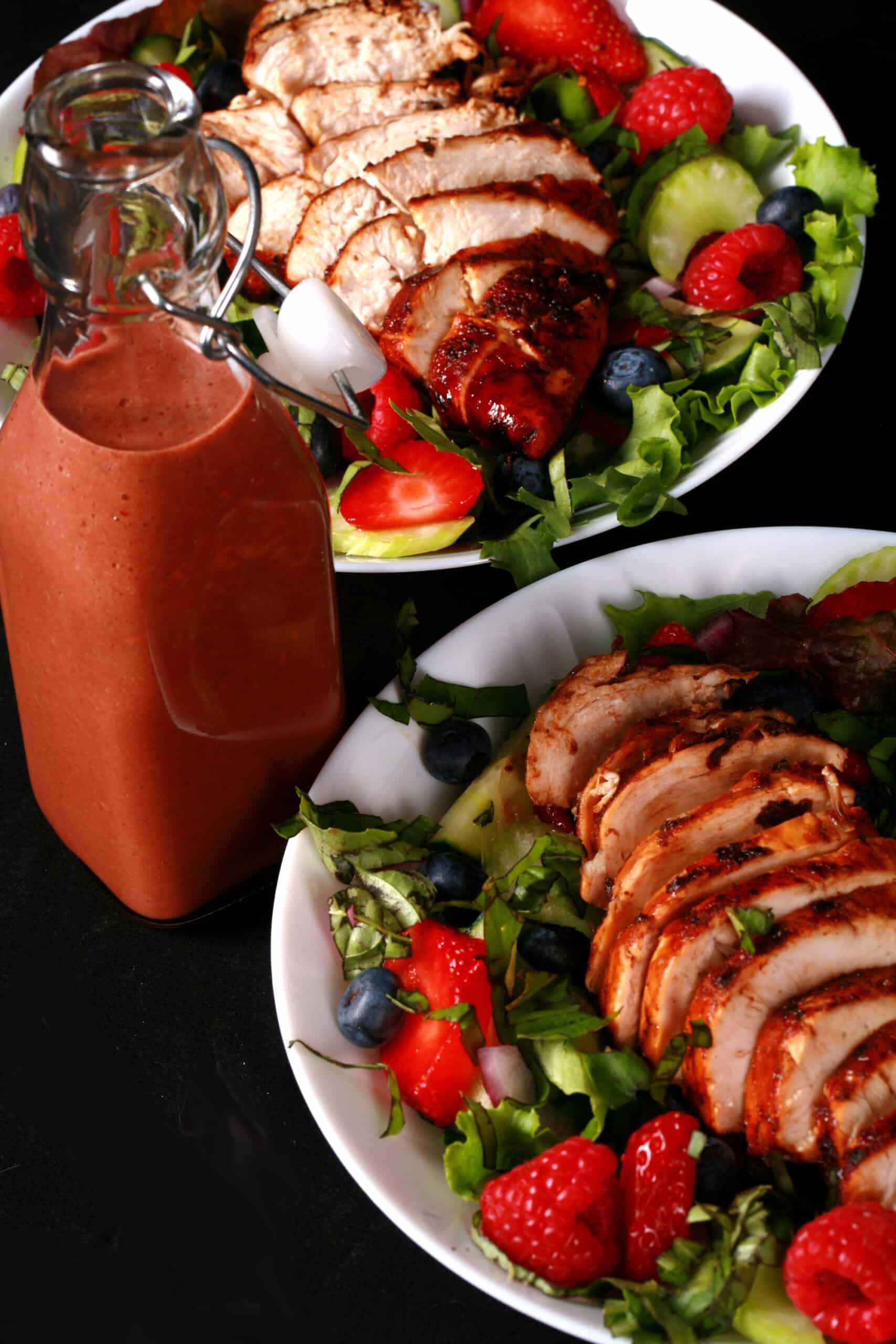 2 summer salads with balsamic glazed chicken and berries. There is a bottle of fresh raspberry balsamic vinaigrette next to it.