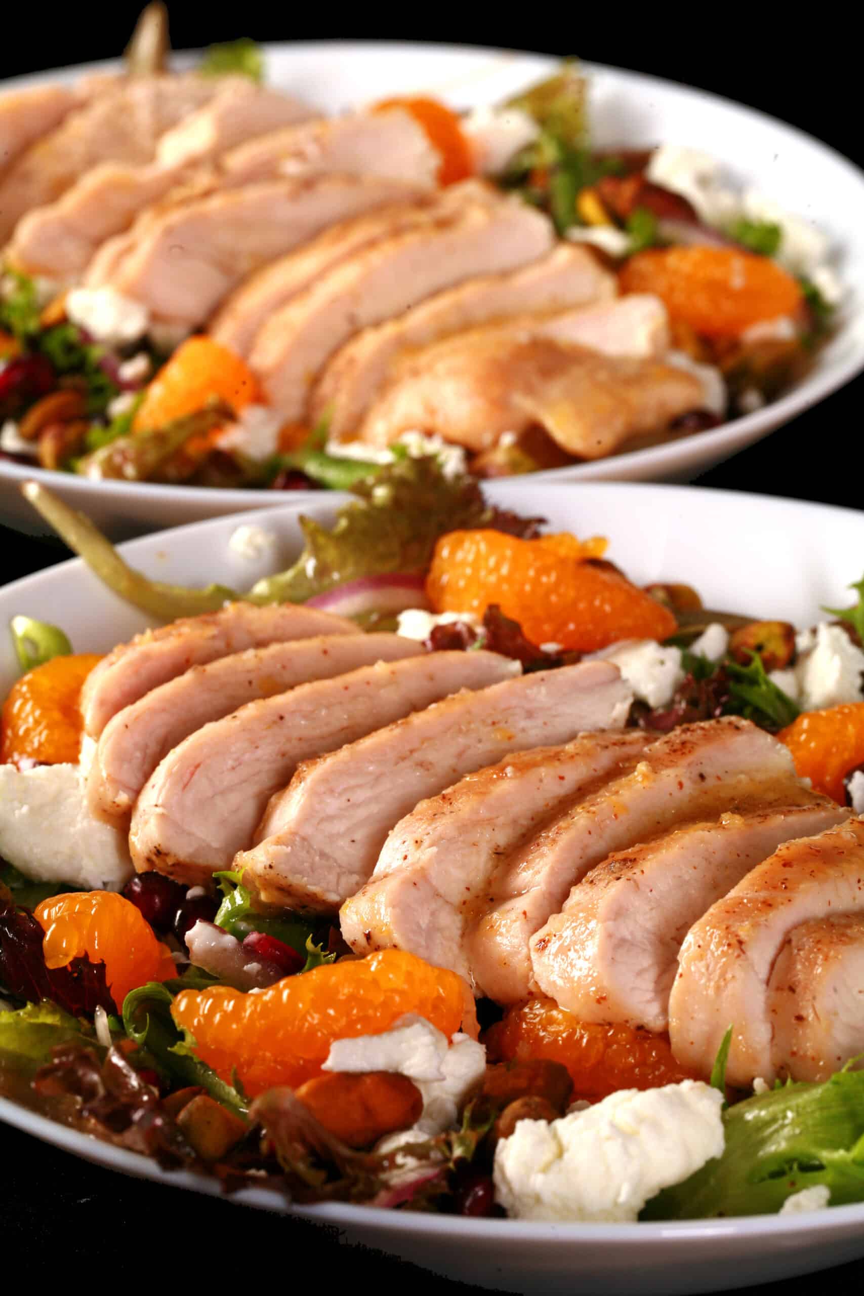 2 bowls of Christmas salad with orange glazed chicken, mandarin orange segments, pomegranates, goat cheese, and pistachios.