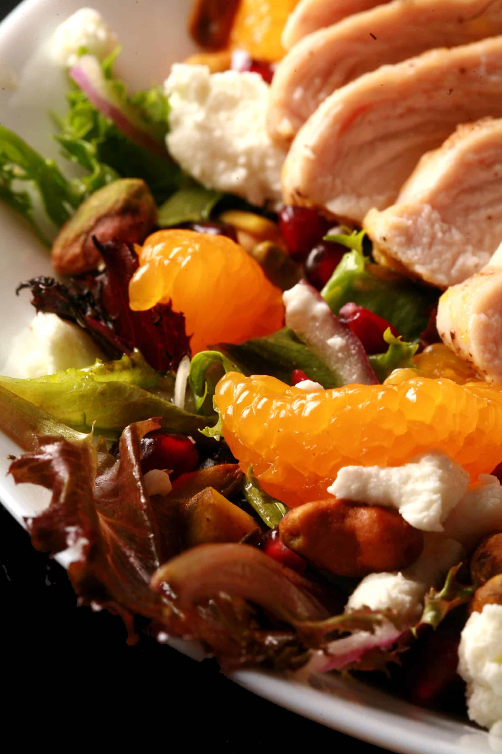 A bowl of Christmas salad with orange glazed chicken, mandarin orange segments, pomegranates, goat cheese, and pistachios.