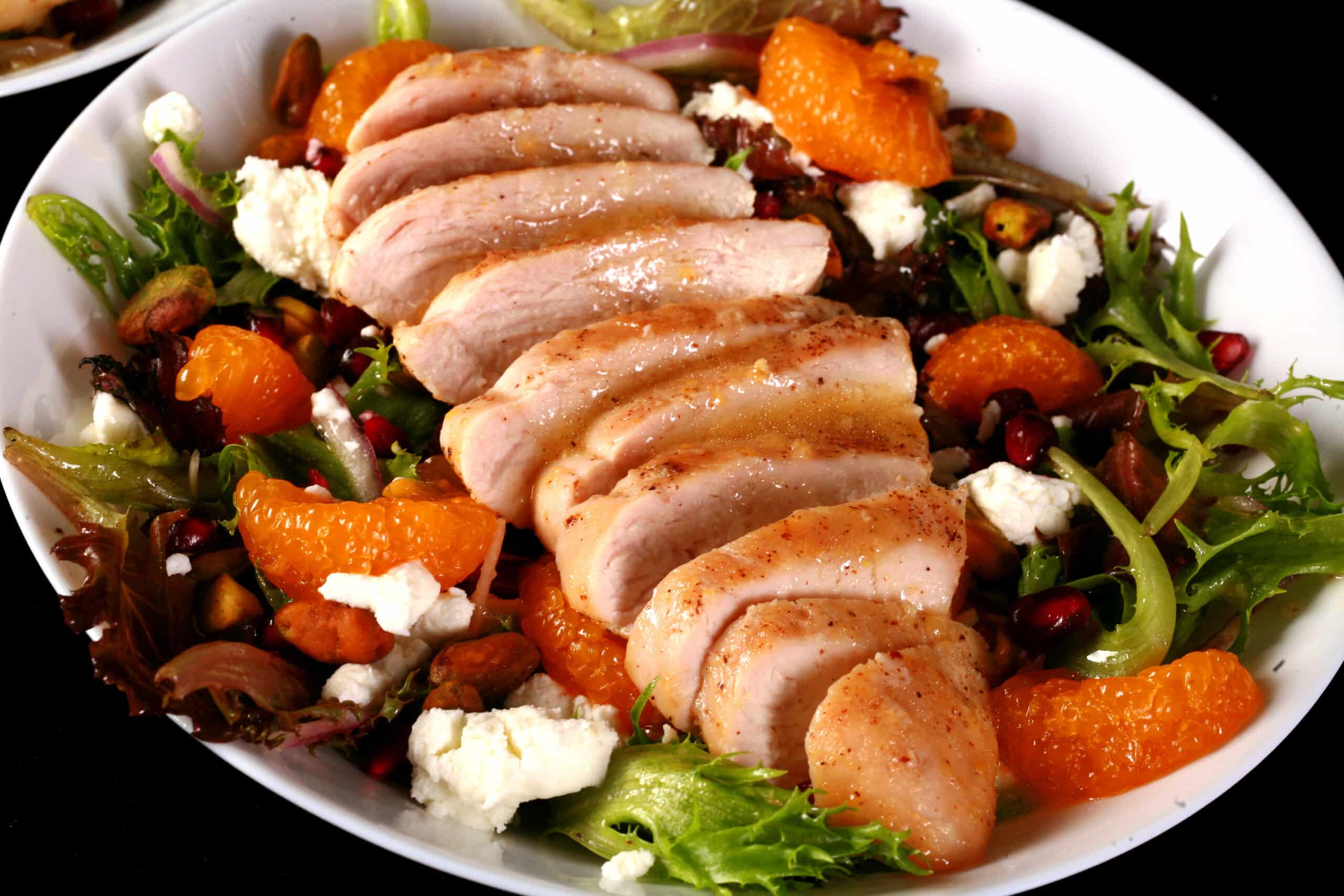 A bowl of Christmas salad with orange glazed chicken, mandarin orange segments, pomegranates, goat cheese, and pistachios.