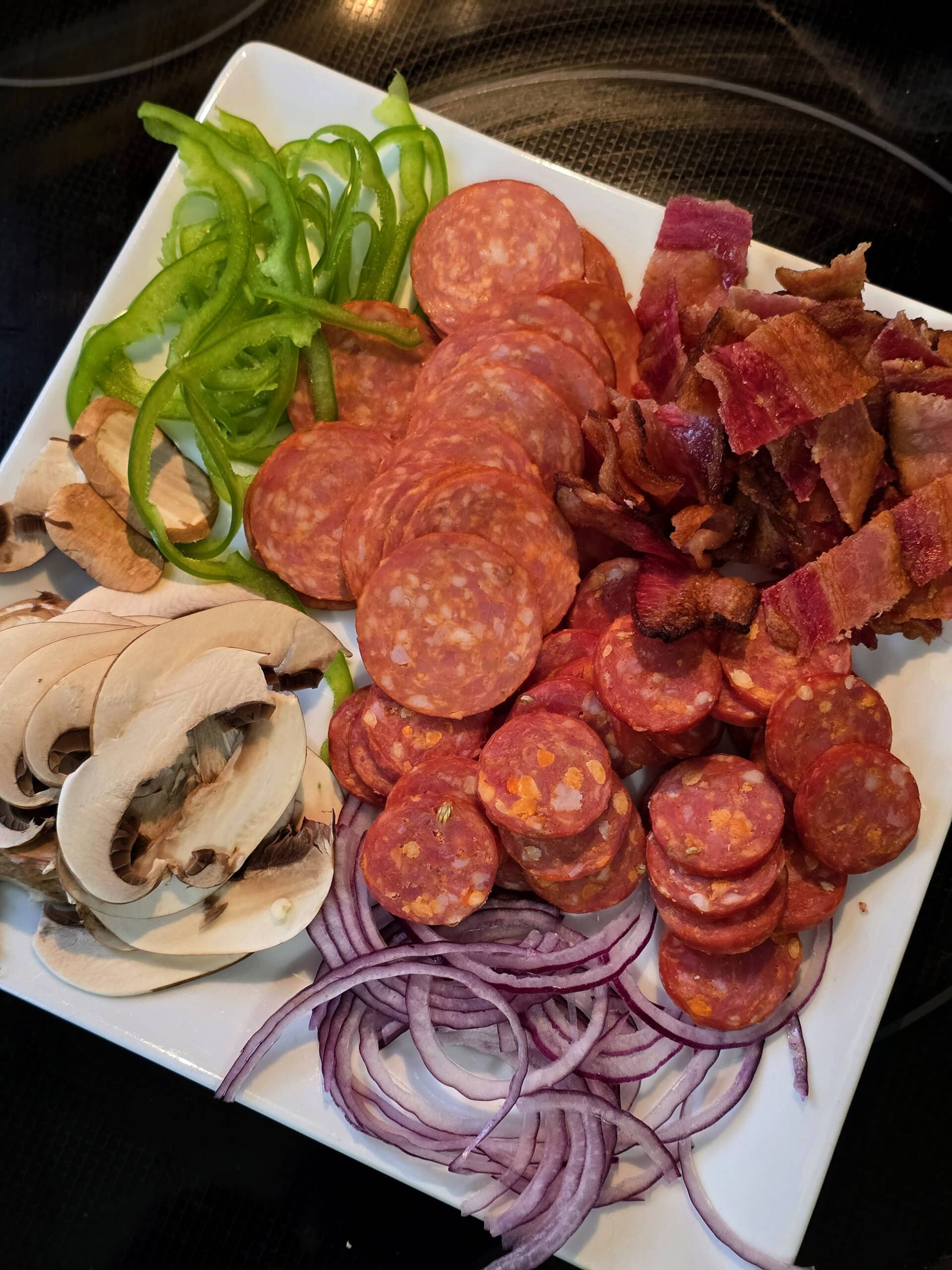 A plate of prepared pizza toppings including pepperoni, bacon, sliced mushrooms, green peppers, and red onions.