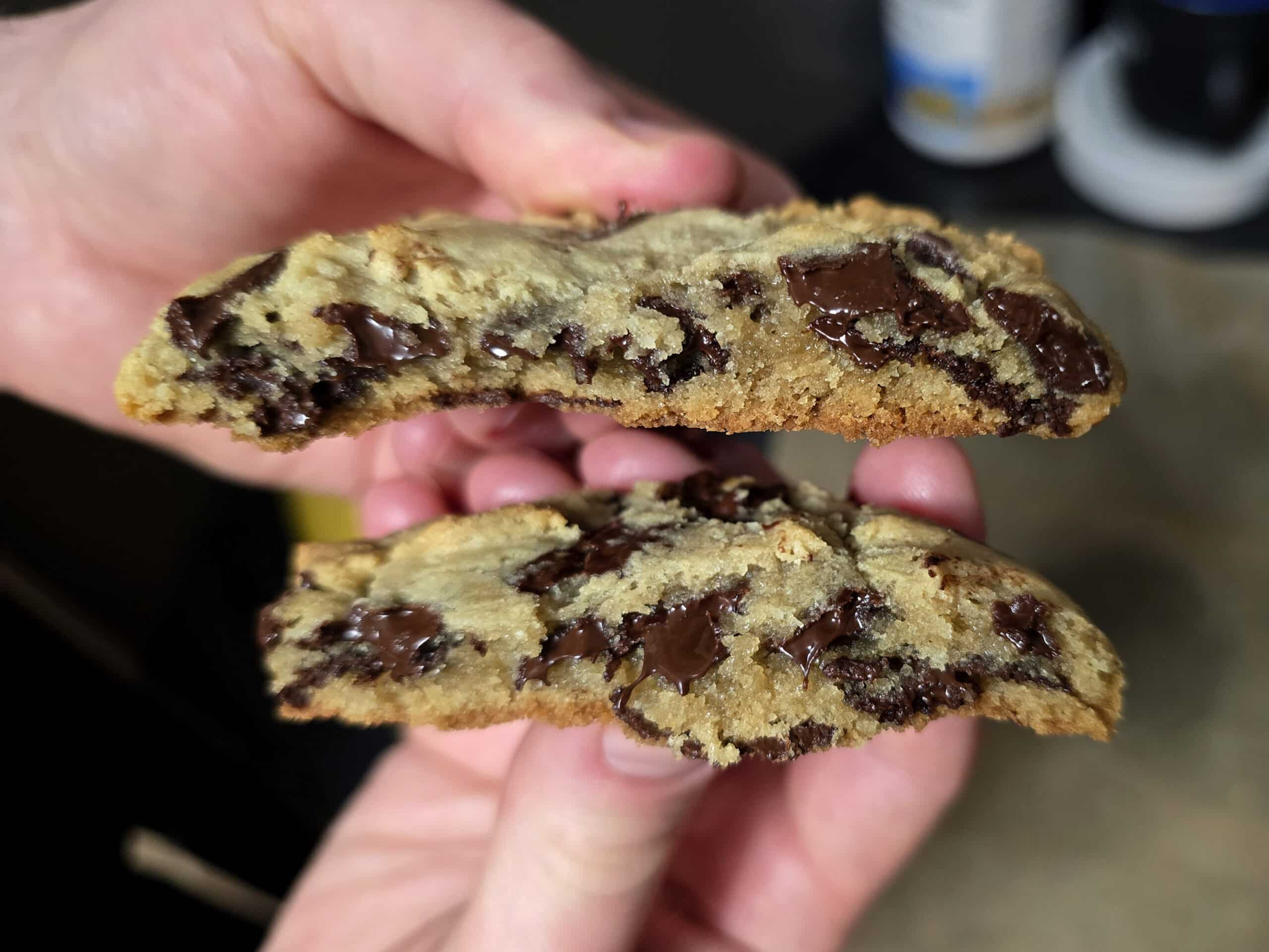 Hands hold a broken in half cookie, with melty chocolate chips.