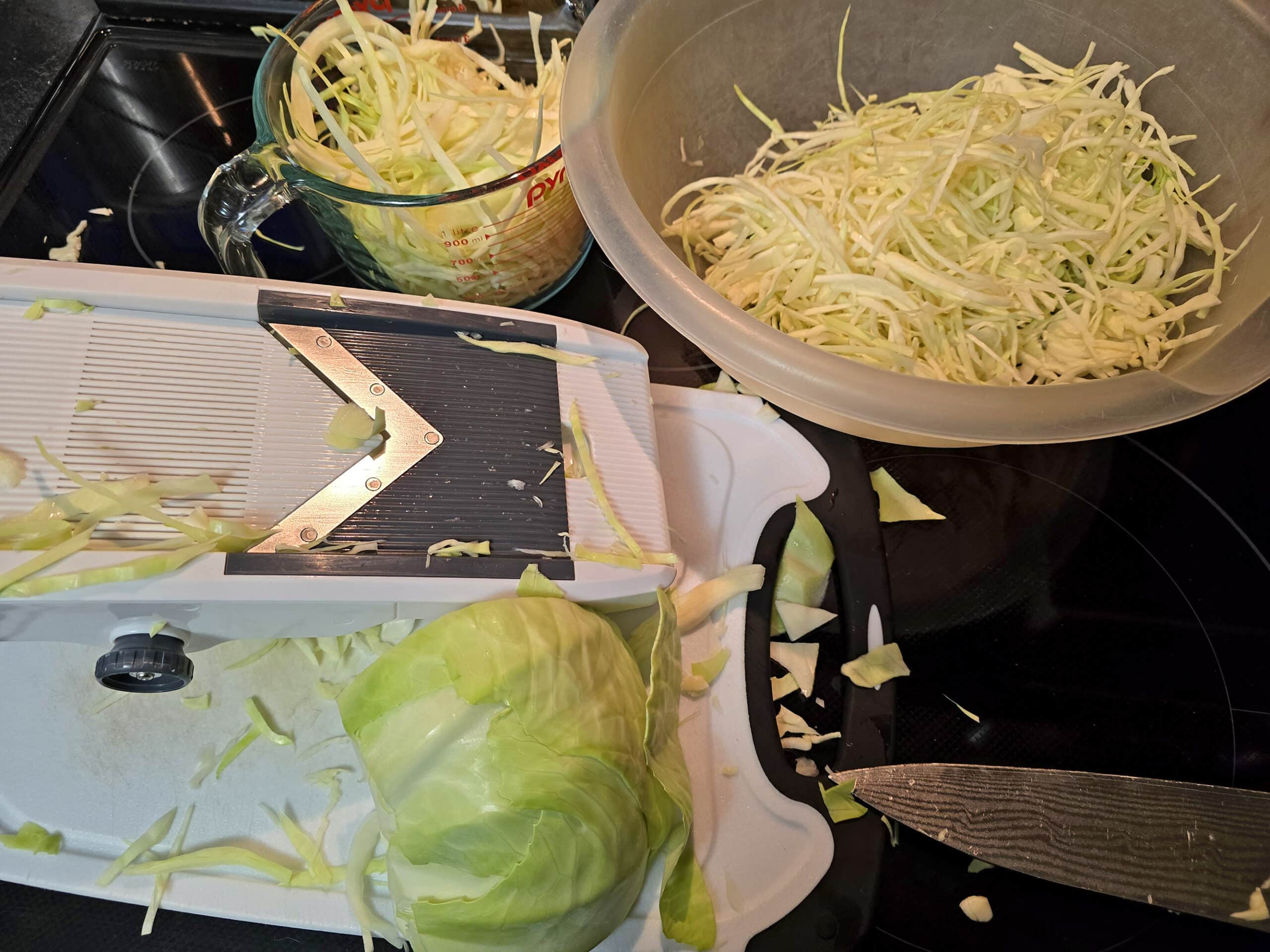 A mandoline slicer being used to shred a green cabbage for coleslaw.