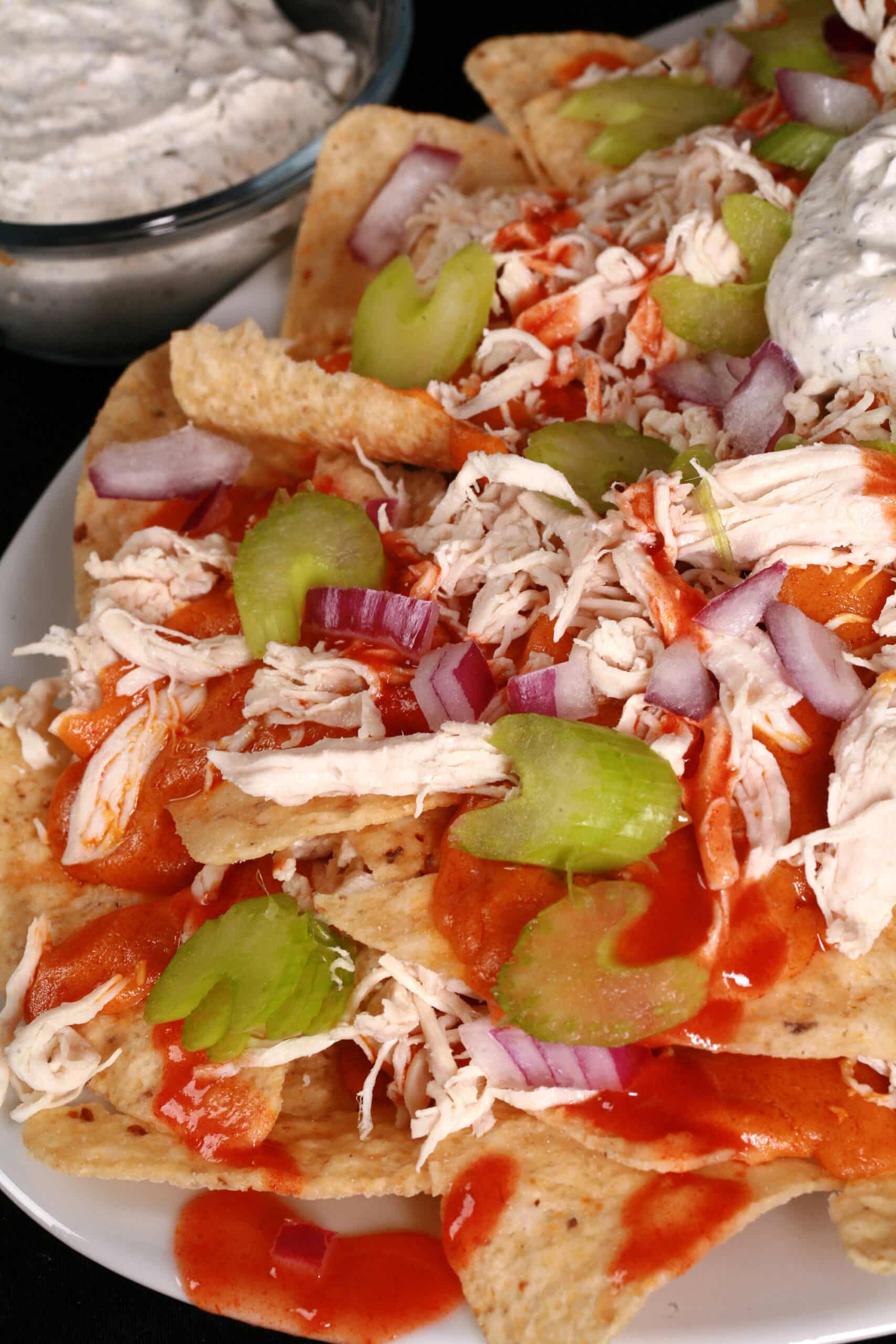 A platter of buffalo chicken nachos, with shredded chicken, Buffalo Wing nacho cheese sauce, hot sauce, red onions, celery, and ranch flavoured sour cream.