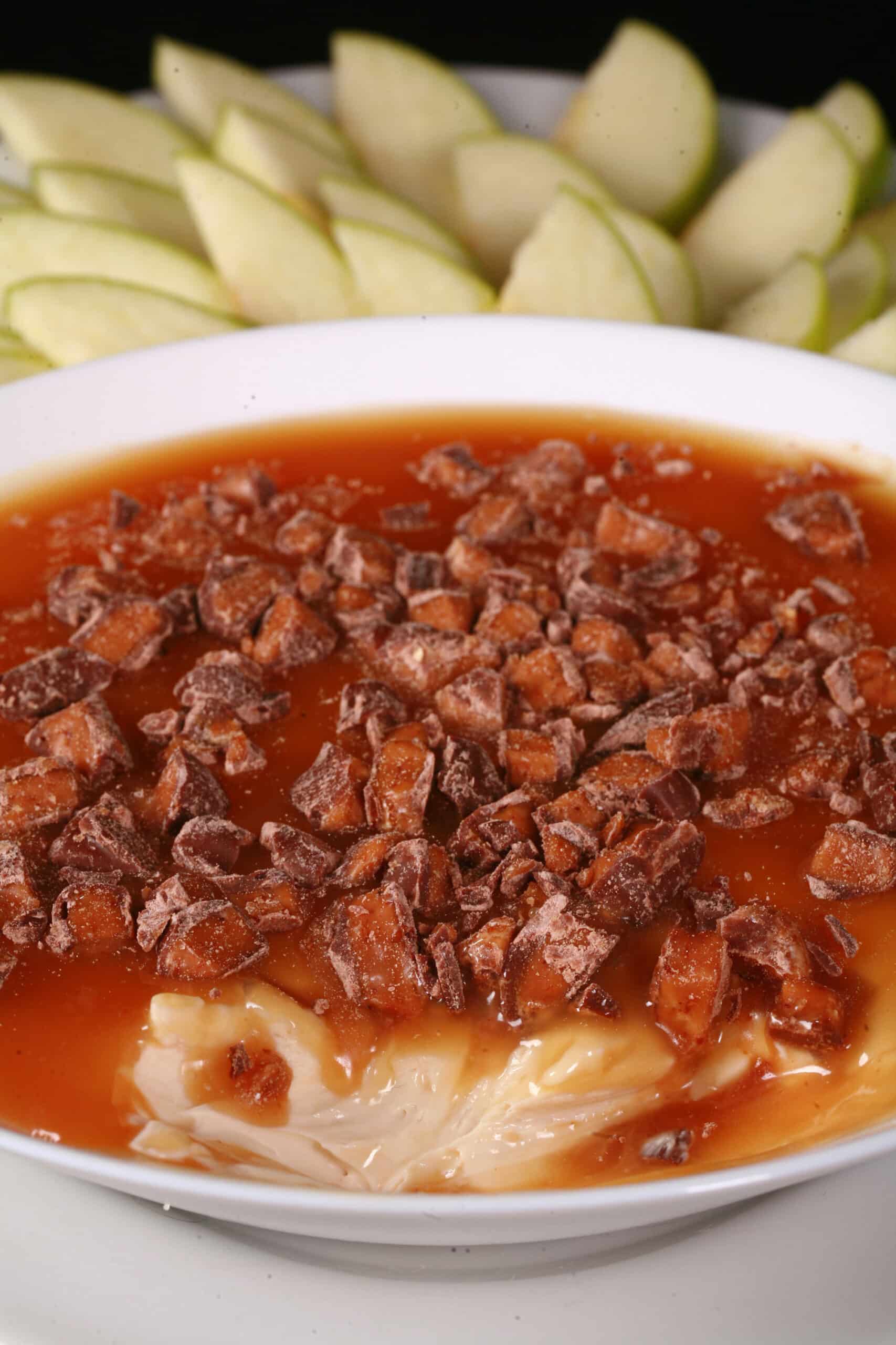 A bowl of caramel apple cheesecake dip, with caramel sauce and chopped Skor bars on top, and sliced granny smith apples behind it.