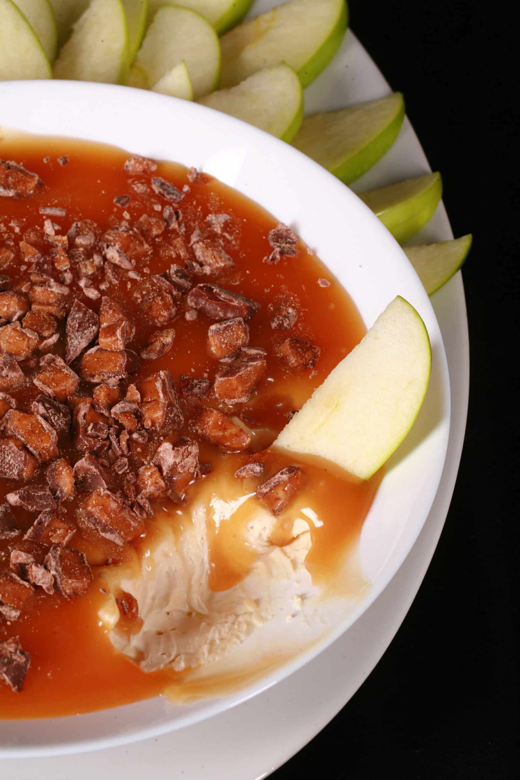 A bowl of cream cheese caramel apple dip, with caramel sauce and chopped Skor bars on top, and sliced granny smith apples behind it.