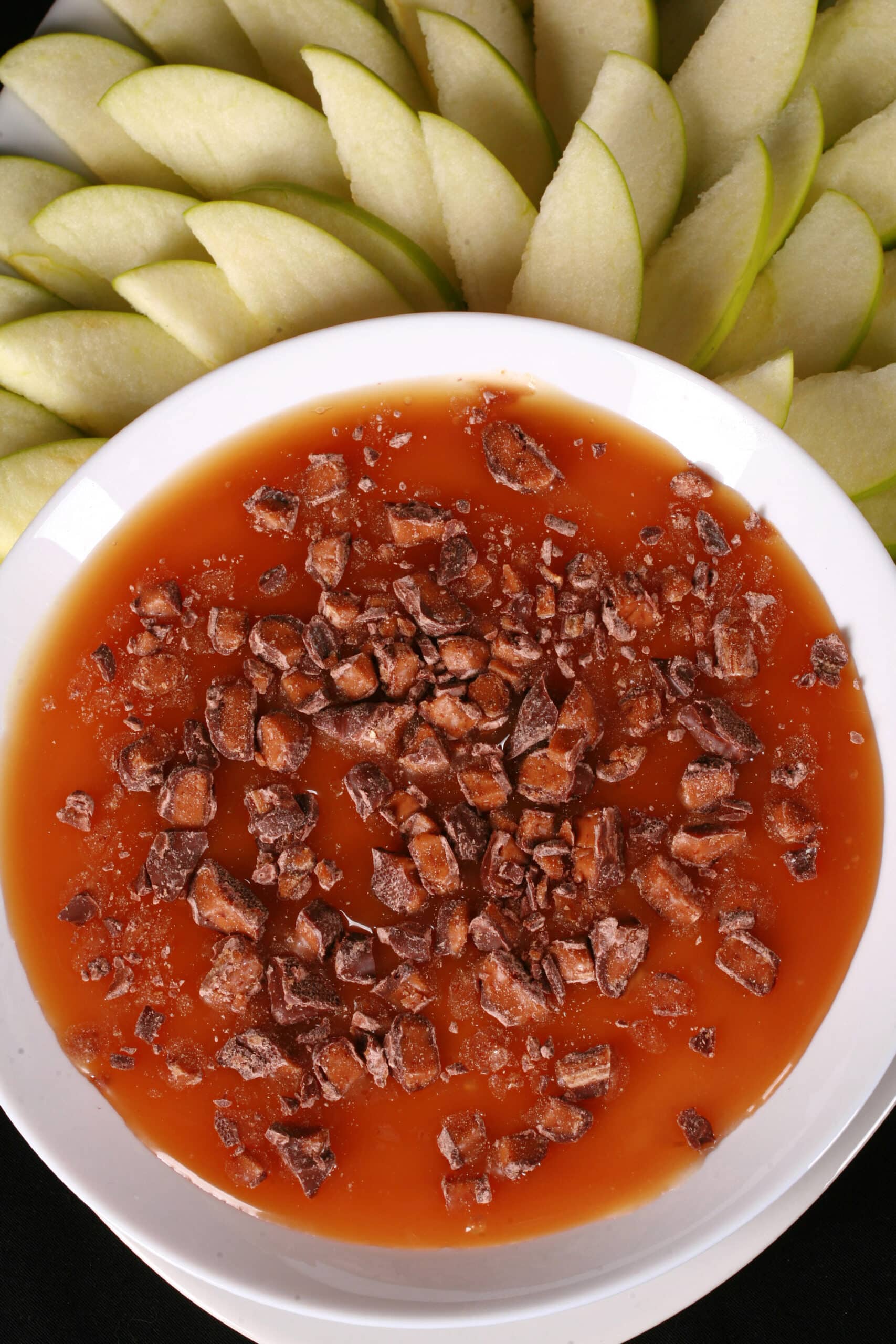 A bowl of caramel apple cheesecake dip, with caramel sauce and chopped Skor bars on top, and sliced granny smith apples behind it.